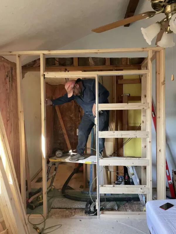 A construction worker builds a wooden framework as part of a primary bath remodel. He is balancing on a work platform while installing wooden studs. The exposed ceiling beams and insulation highlight the major renovation work in progress.