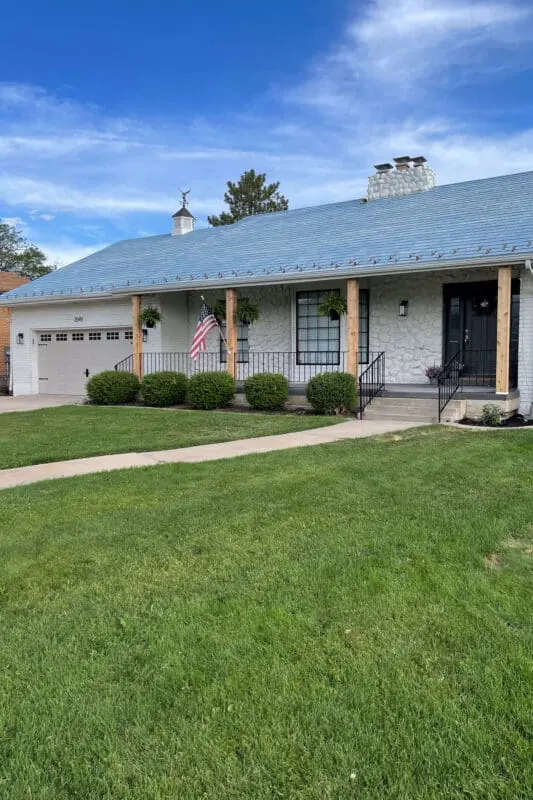 White painted brick house with natural wood porch column wraps and stone facade, styled with hanging ferns and an American flag for a cozy, classic curb appeal.