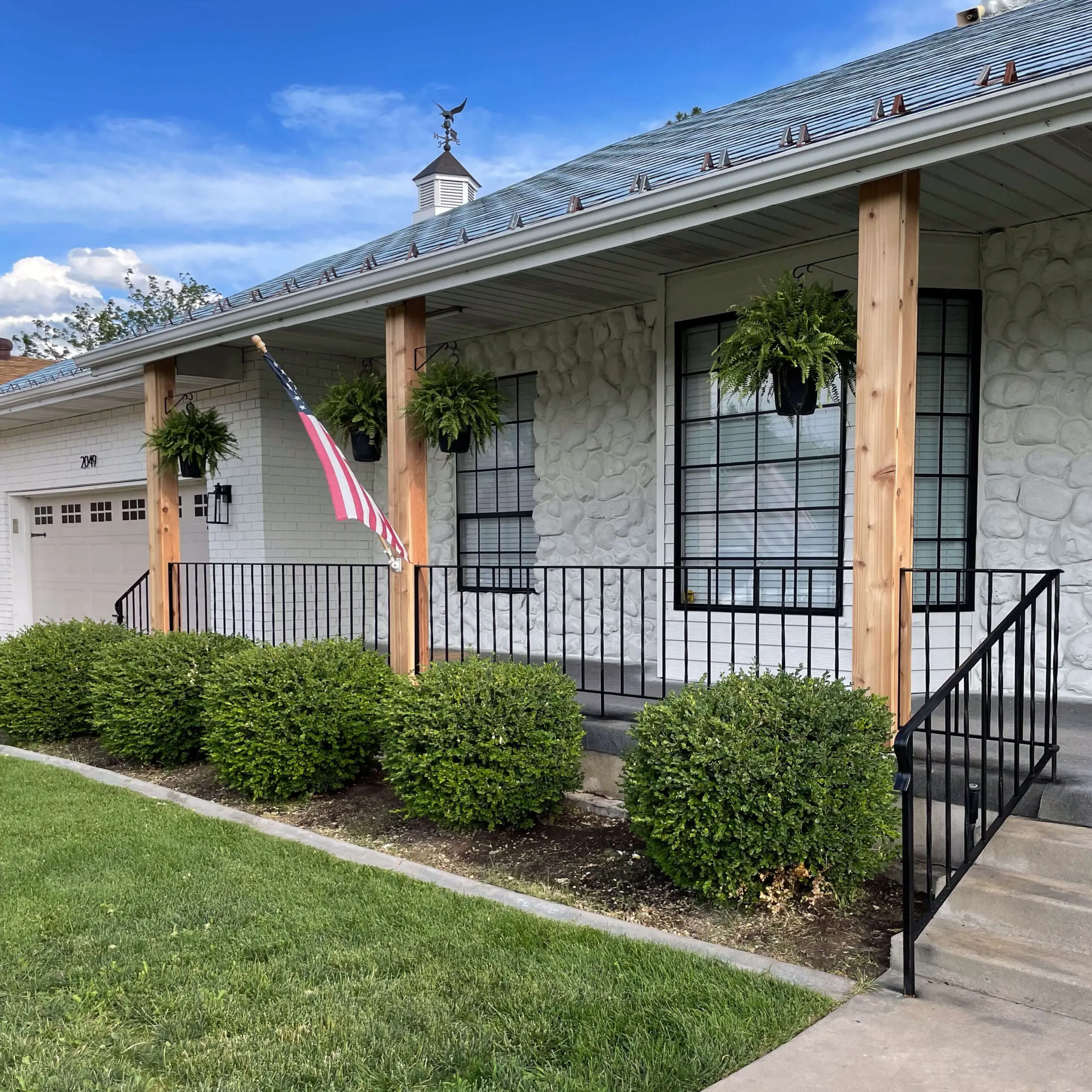Angled view of front porch showcasing freshly installed wood porch column wraps, black railing, manicured hedges, and a patriotic flag display.