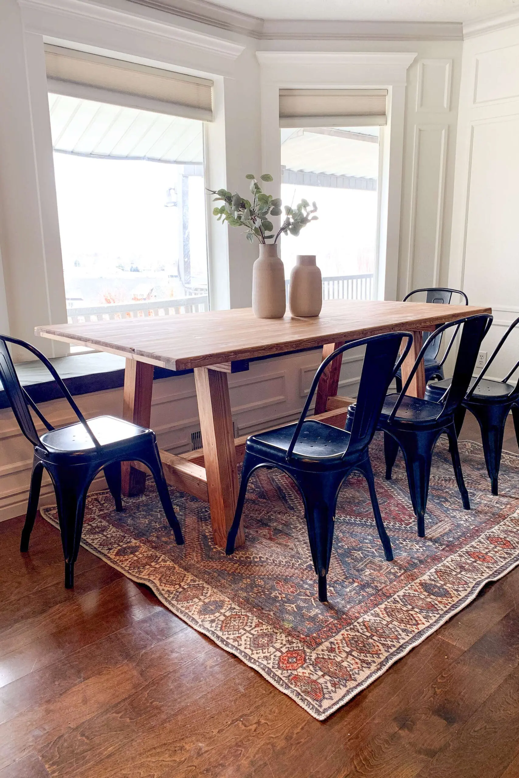 Cozy dining area featuring a DIY farmhouse table with black metal chairs, styled with vases and placed on a patterned rug.