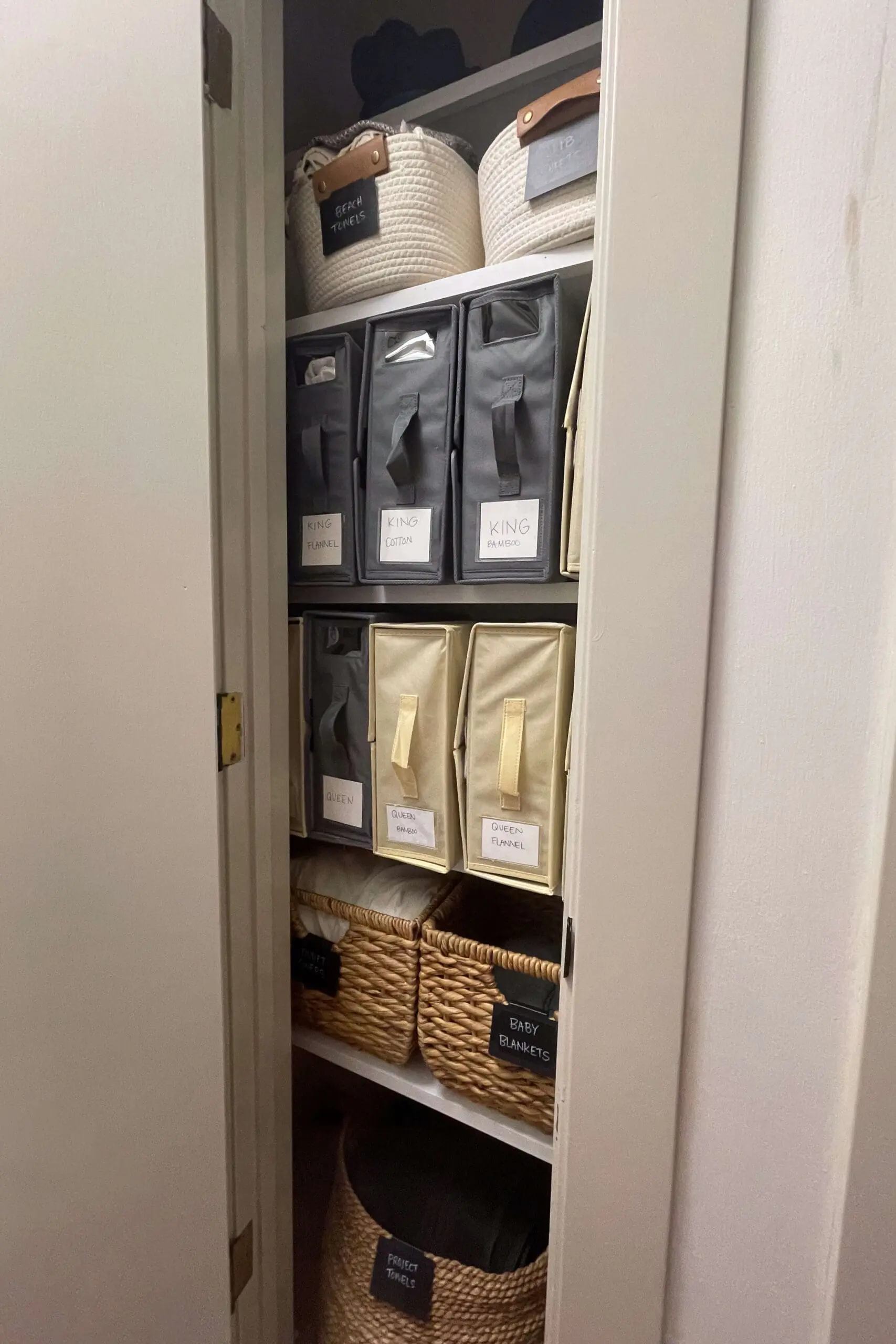 Neatly organized linen closet with labeled fabric storage bins and woven baskets for easy access to bedding and towels.