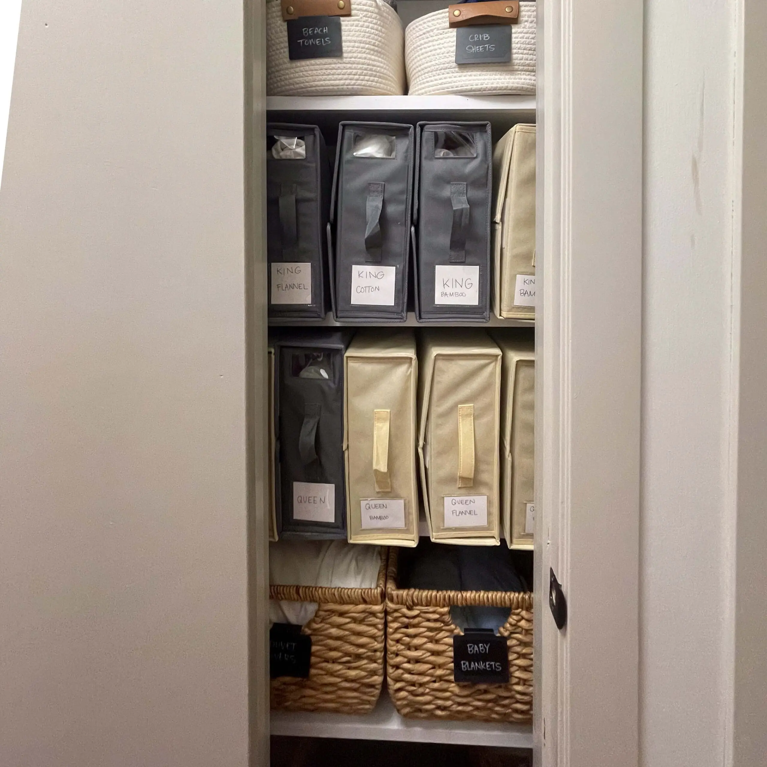 Linen closet organization with labeled storage bins for king and queen sheets, crib sheets, and beach towels in woven and fabric storage containers.