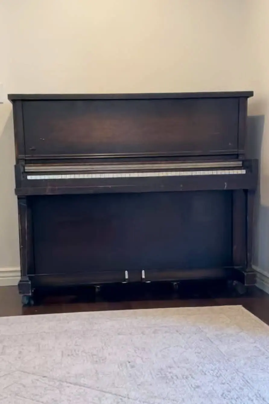 Vintage dark brown upright piano in its original worn finish, positioned against a neutral wall, sitting on a dark hardwood floor with a light-colored rug in front.
