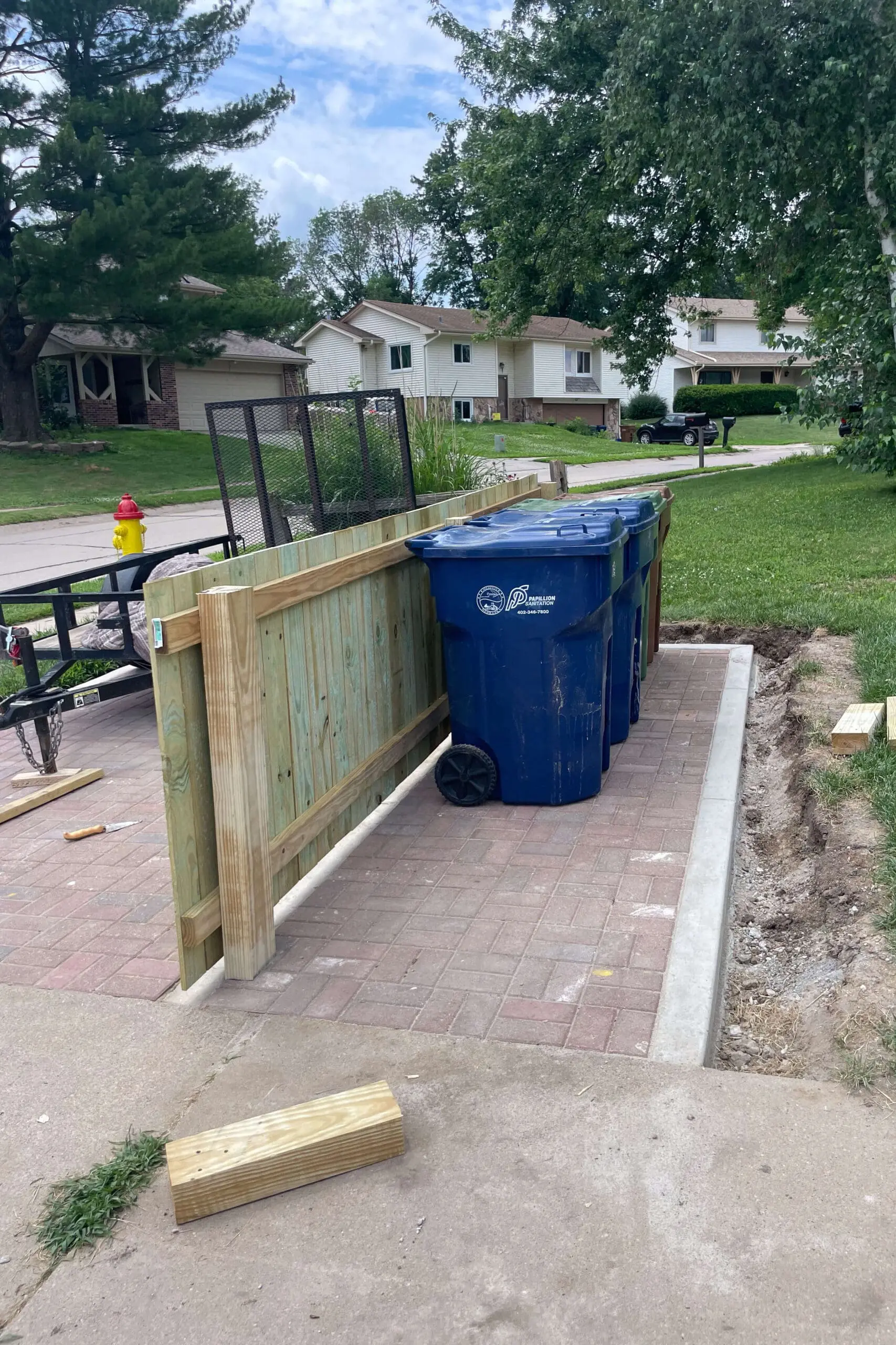 Custom-built wooden privacy fence hides trash bins on a paved brick pad for a clean, organized curbside look.