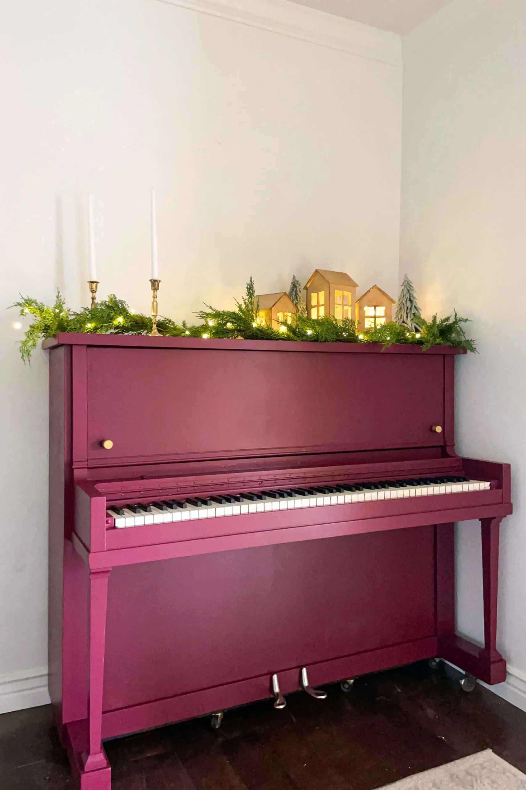A beautifully painted burgundy upright piano styled for the holidays with warm glowing wooden houses, fresh greenery, fairy lights, and brass candlesticks on top.