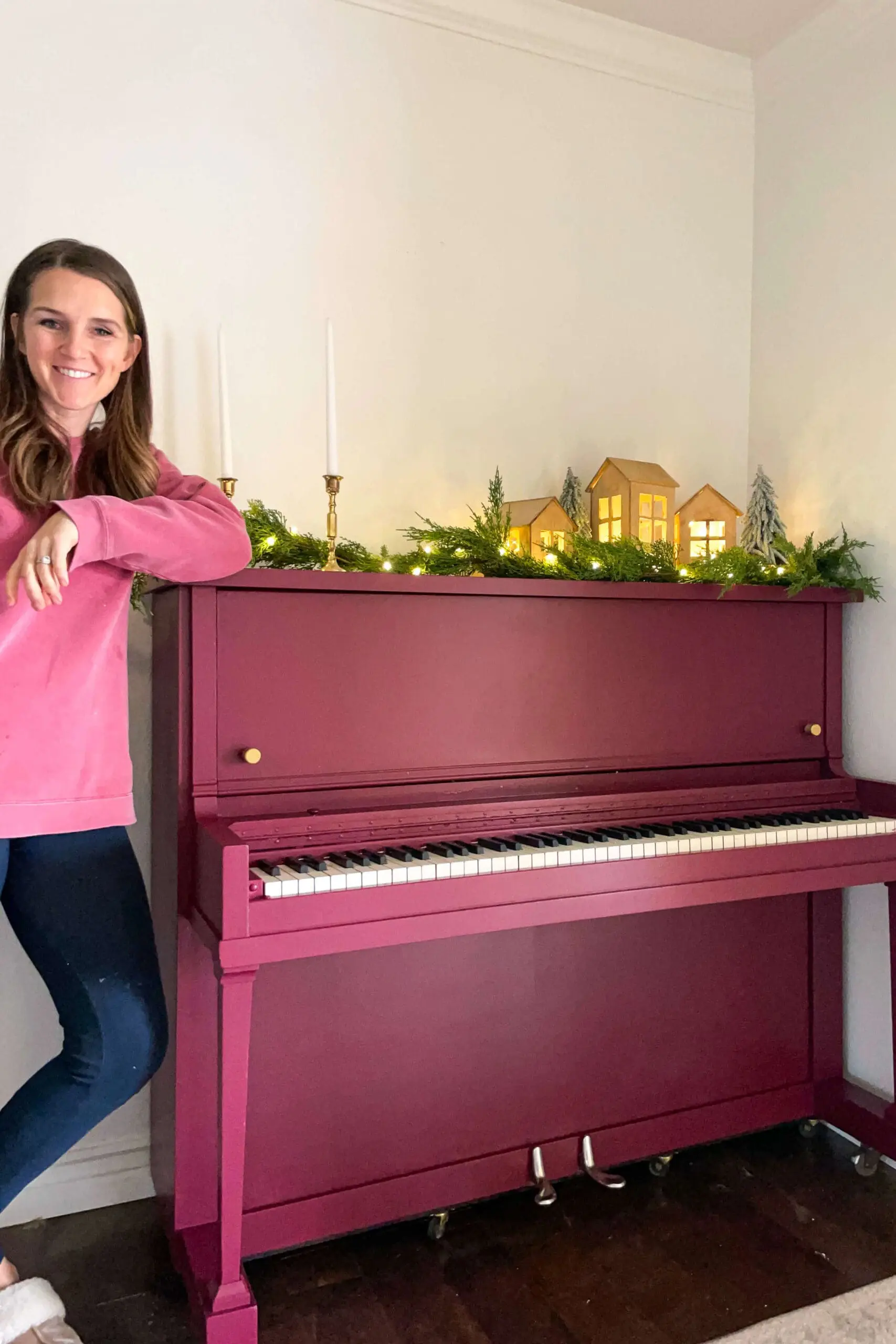 Woman smiling next to her freshly painted burgundy upright piano, decorated with festive Christmas greenery, glowing wooden houses, and brass candlesticks.