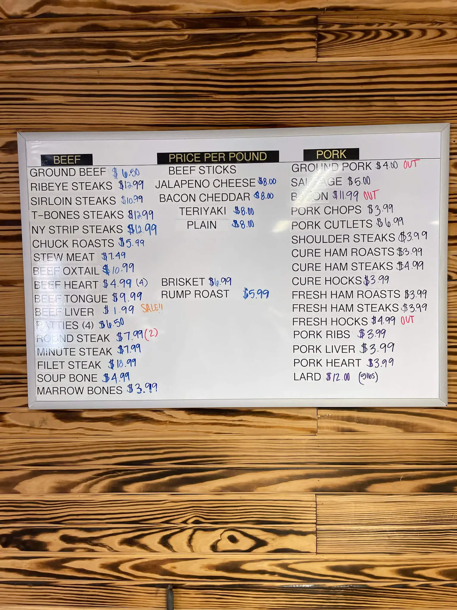 A whiteboard menu at a local butcher shop displaying prices for beef and pork cuts, ideal for a carnivore diet grocery haul.