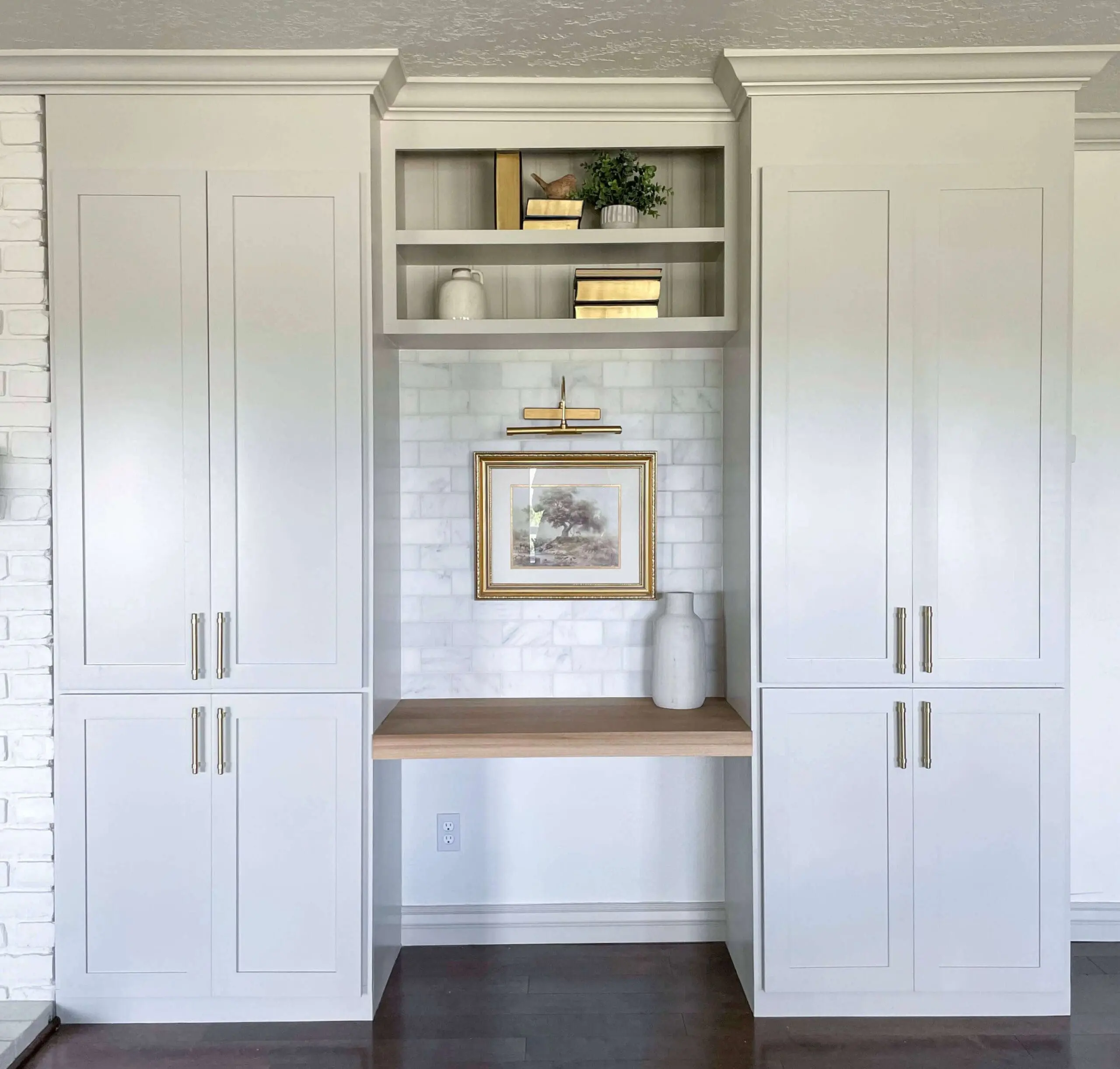 Finished built-in office nook with a wooden desk, white shaker cabinets, gold hardware, and a marble backsplash.