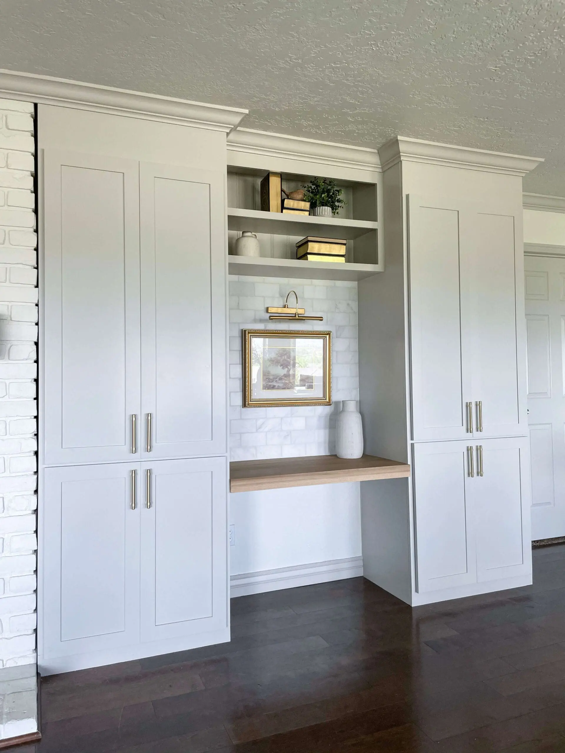 A set of built-in cabinets with shaker-style doors, gold handles, and a light wood floating desk, styled with books, greenery, and decor.