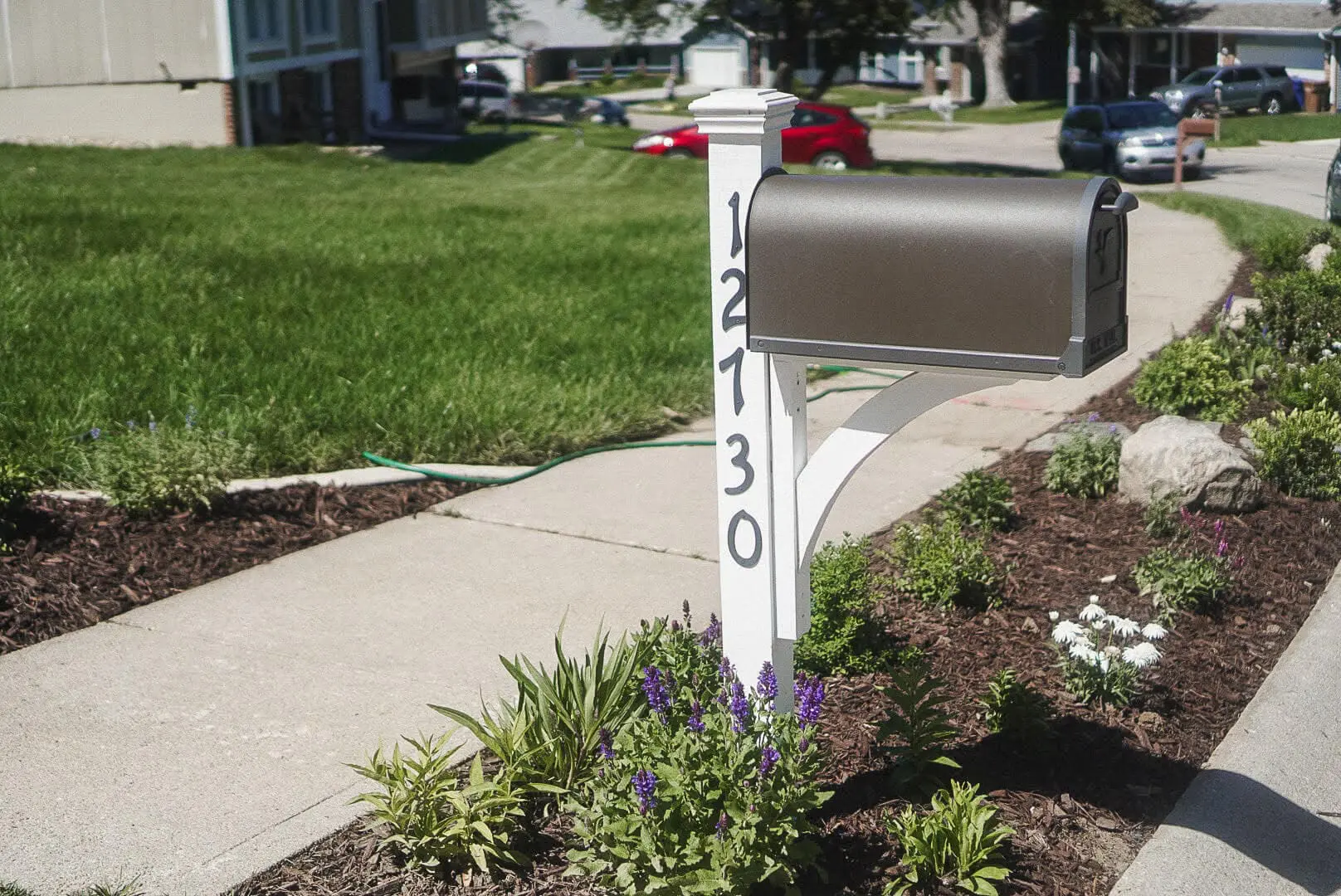 DIY built mailbox with black metal box mounted on a crisp white post, featuring bold house numbers painted vertically. Surrounding landscaping includes blooming white daisies, purple salvia, and mulch beds bordered by concrete edging. The fresh curb appeal is all part of a DIY exterior house remodel.