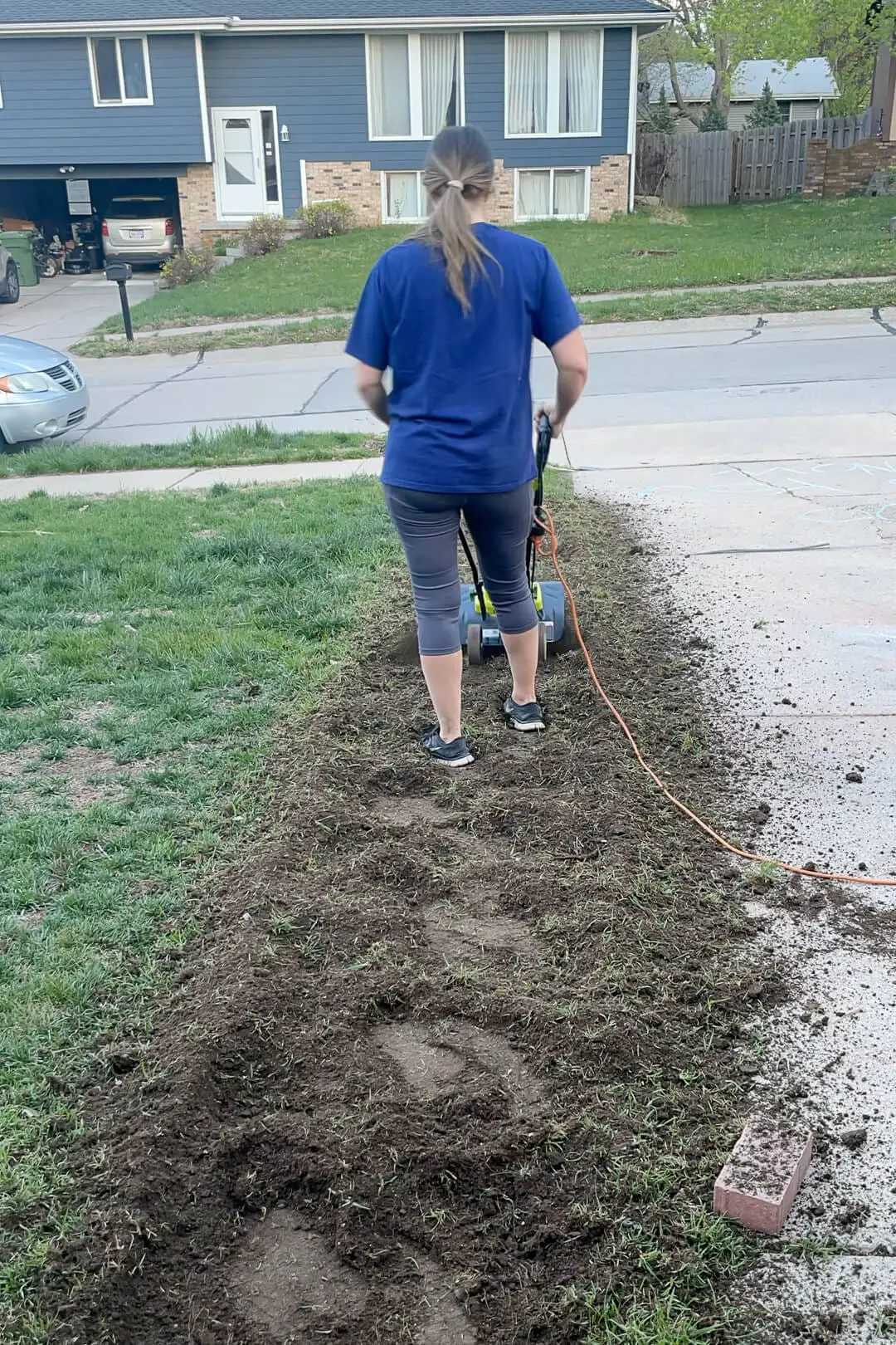 Woman using an electric tiller to break up a strip of soil along the edge of a driveway. She’s working in workout clothes, prepping the area for a new sidewalk, with patchy grass and a quiet suburban street in the background.