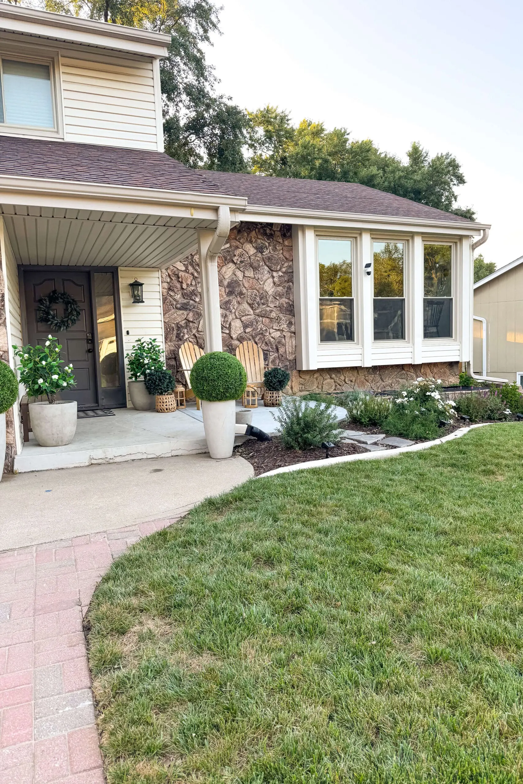 Refreshed front porch with modern black door, decorative wreath, and oversized planters filled with greenery. New curved concrete edging frames a mulched garden bed with lush green shrubs, white flowers, and a stone path leading from the lawn. Landscaping made a big difference in this exterior house remodel.