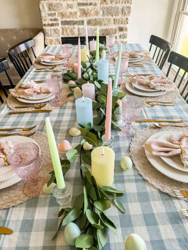 Overhead view of Easter table decor with pastel taper and pillar candles, eucalyptus greenery, speckled eggs, and pink vintage goblets on a blue gingham tablecloth.