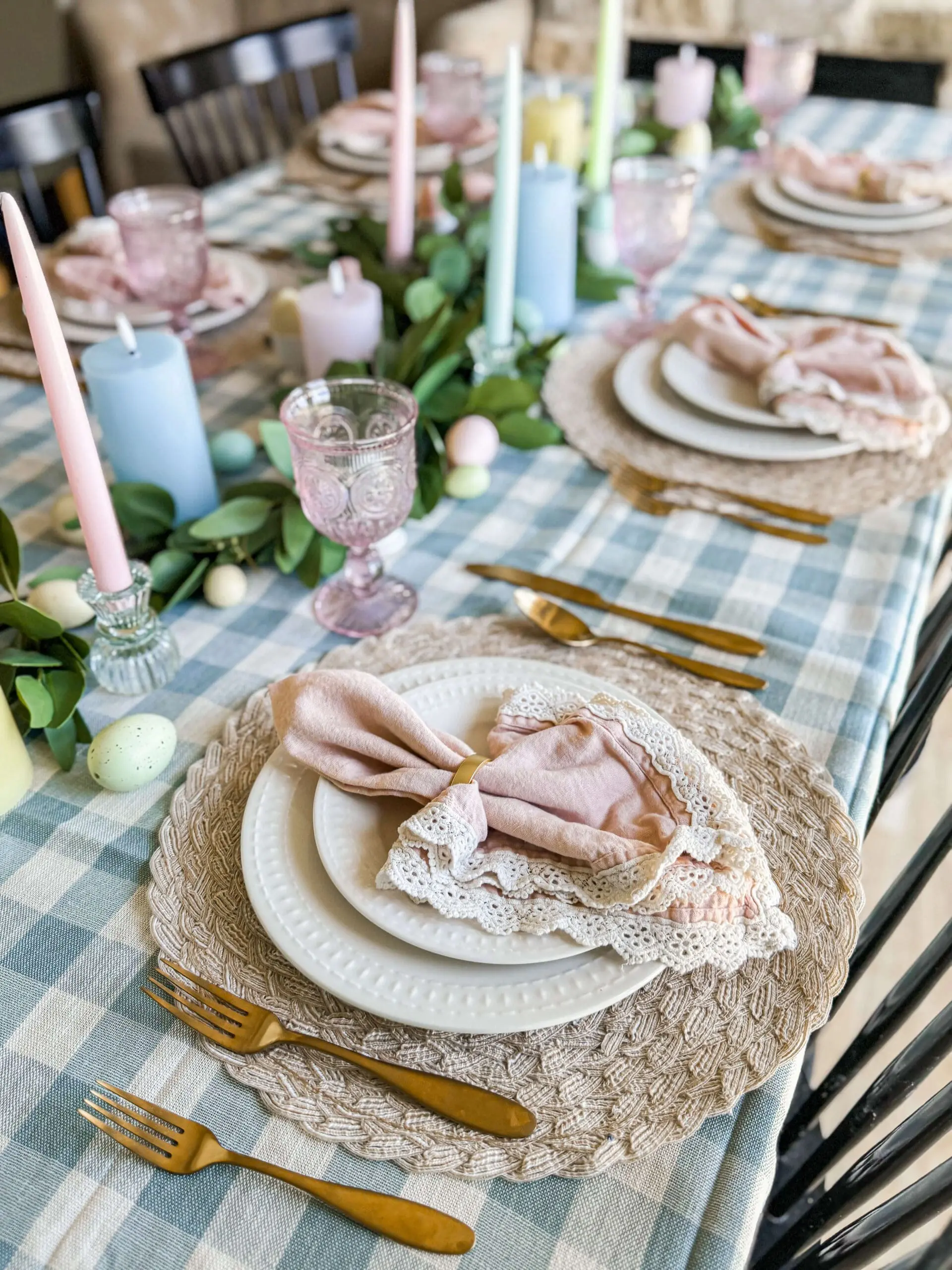 Full view of spring Easter table decor with a blue gingham tablecloth, pastel taper and pillar candles, pink vintage-style glassware, eucalyptus garland, speckled eggs, and layered woven placemats topped with lace-trimmed napkins.