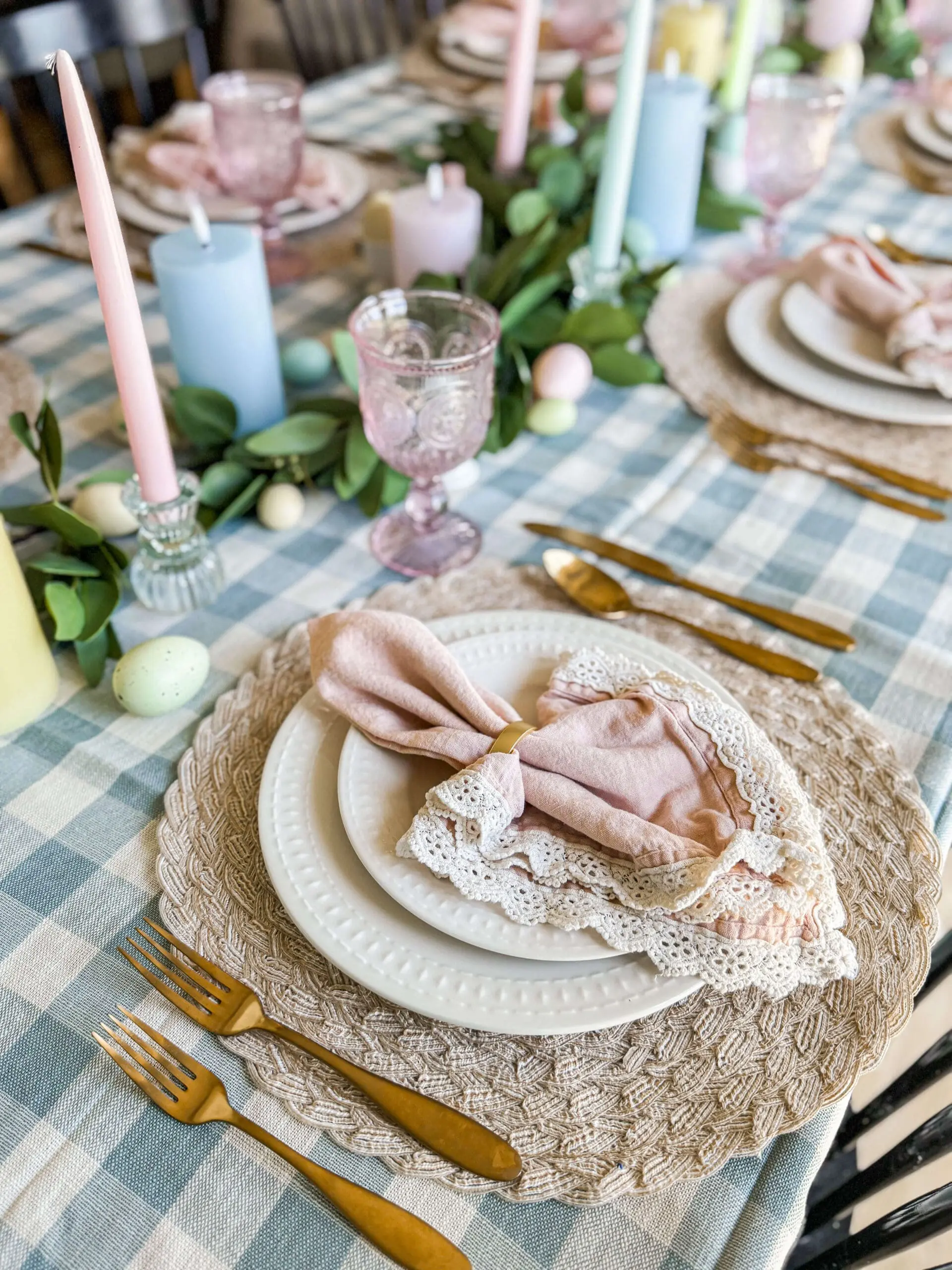 Easter table decor with soft pastel candles, woven placemats, lace napkins, and gold flatware styled on a blue gingham tablecloth