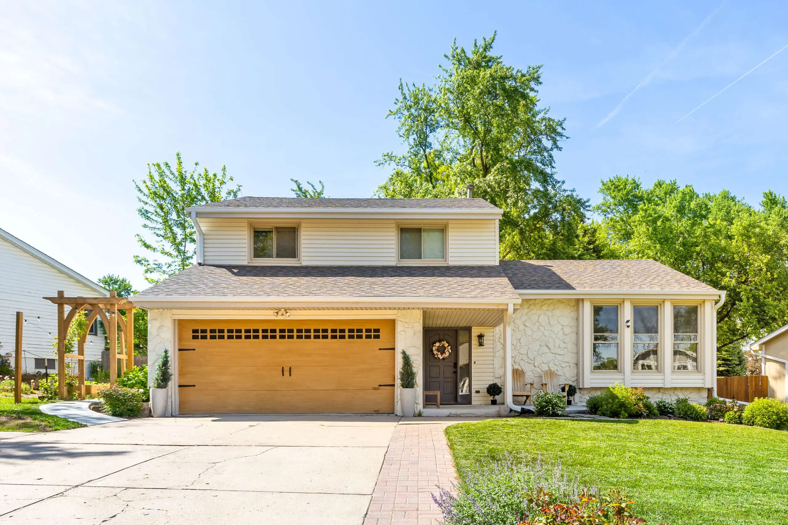 Front view of a freshly updated home exterior featuring a warm wood-tone garage door, stone facade, gray front door, and a wooden pergola with garden landscaping.