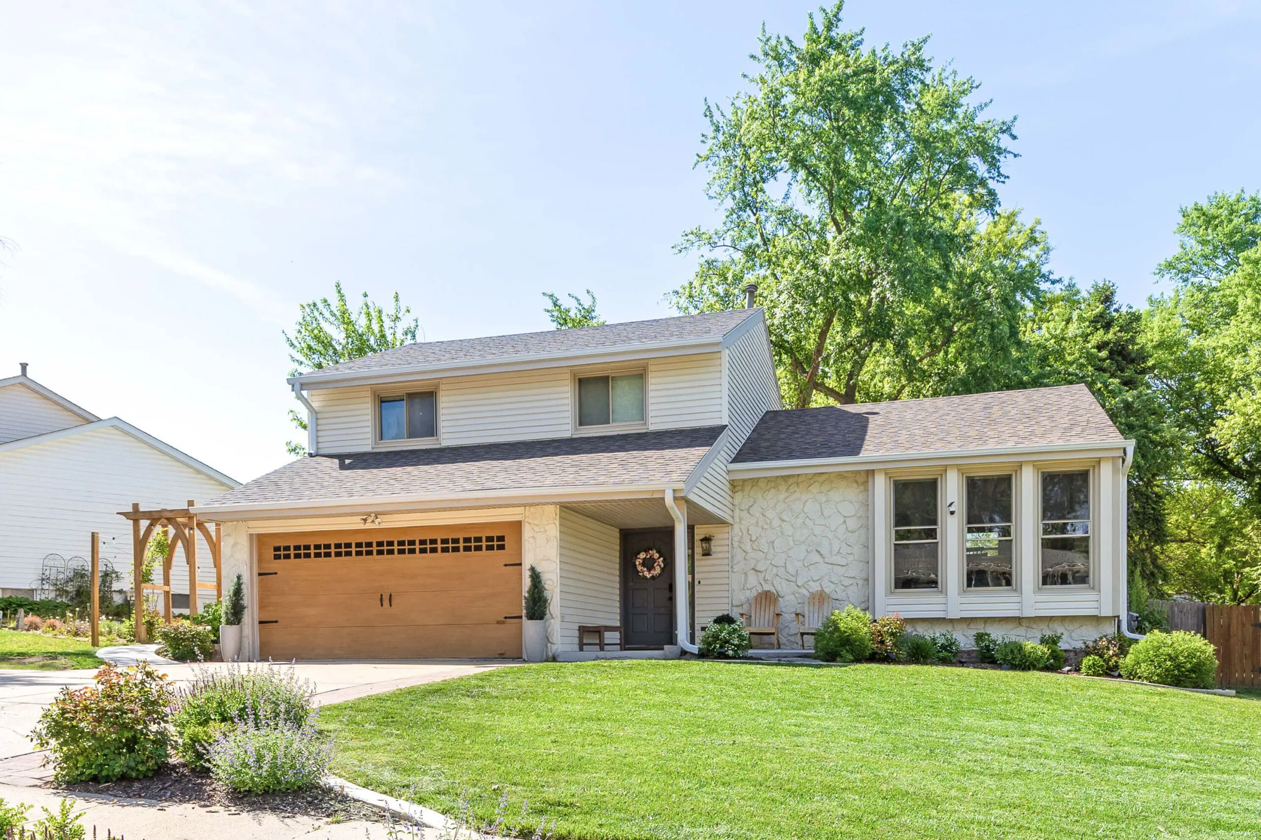Front view of a freshly updated home exterior featuring a warm wood-tone garage door, stone facade, gray front door, and a wooden pergola with garden landscaping.