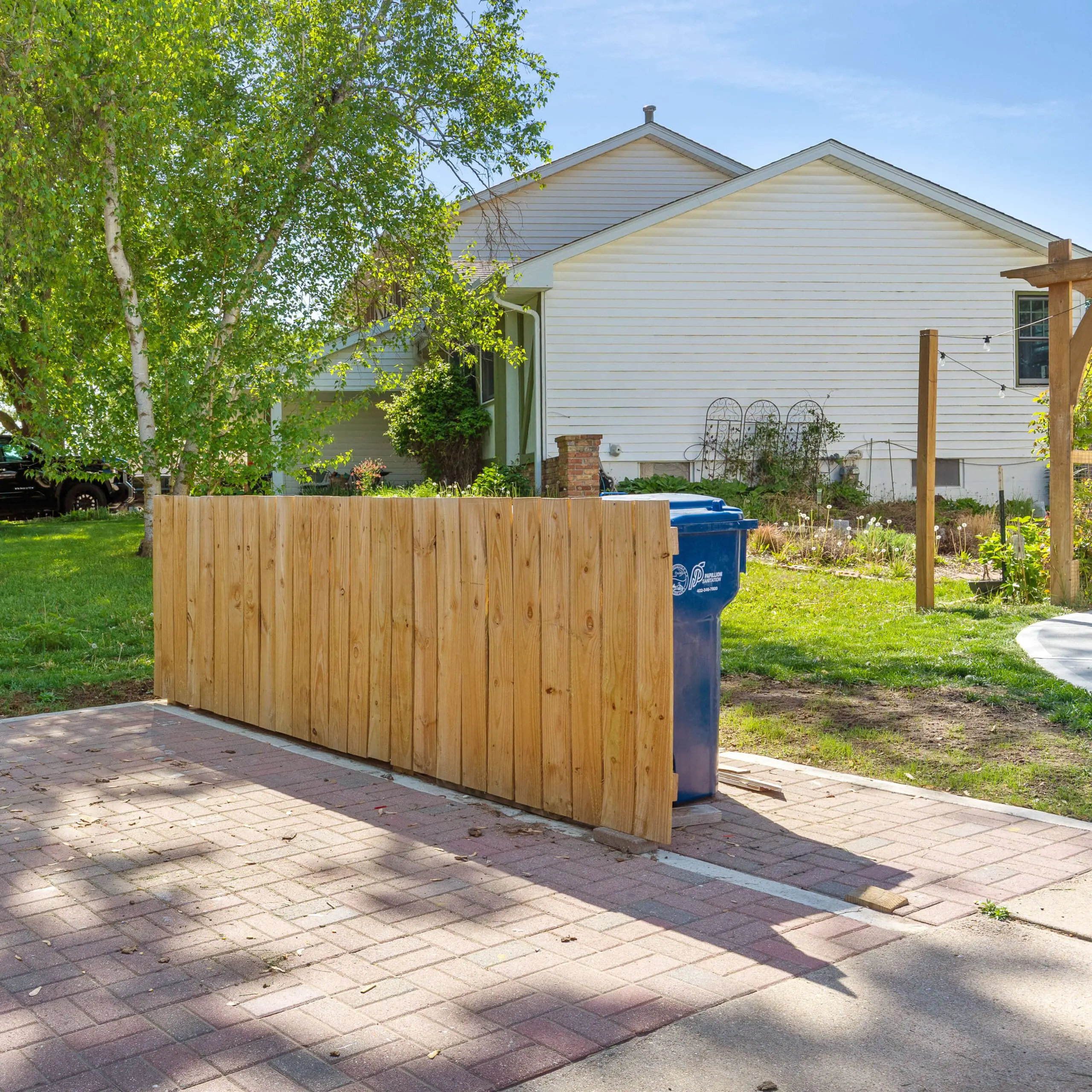 Side angle of a wooden trash enclosure hiding a blue bin, installed over a red brick paver pad with surrounding trees and garden elements in the background.