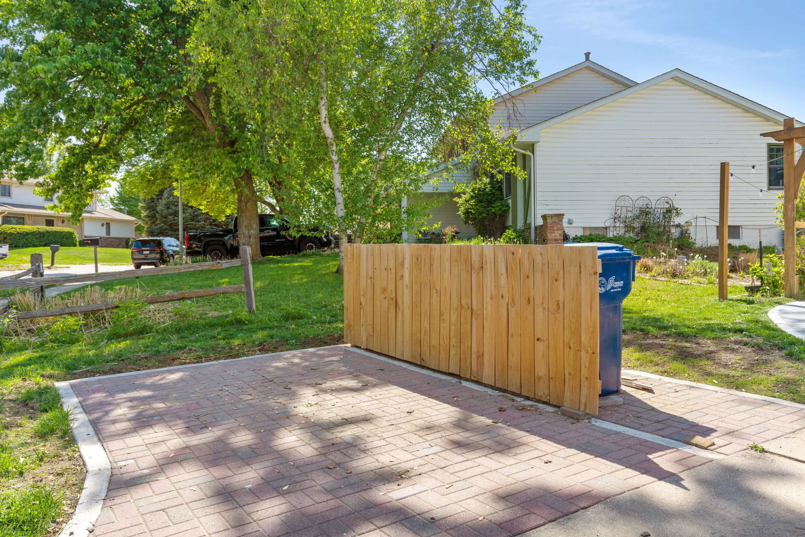 Side angle of a wooden trash enclosure hiding a blue bin, installed over a red brick paver pad with surrounding trees and garden elements in the background.


