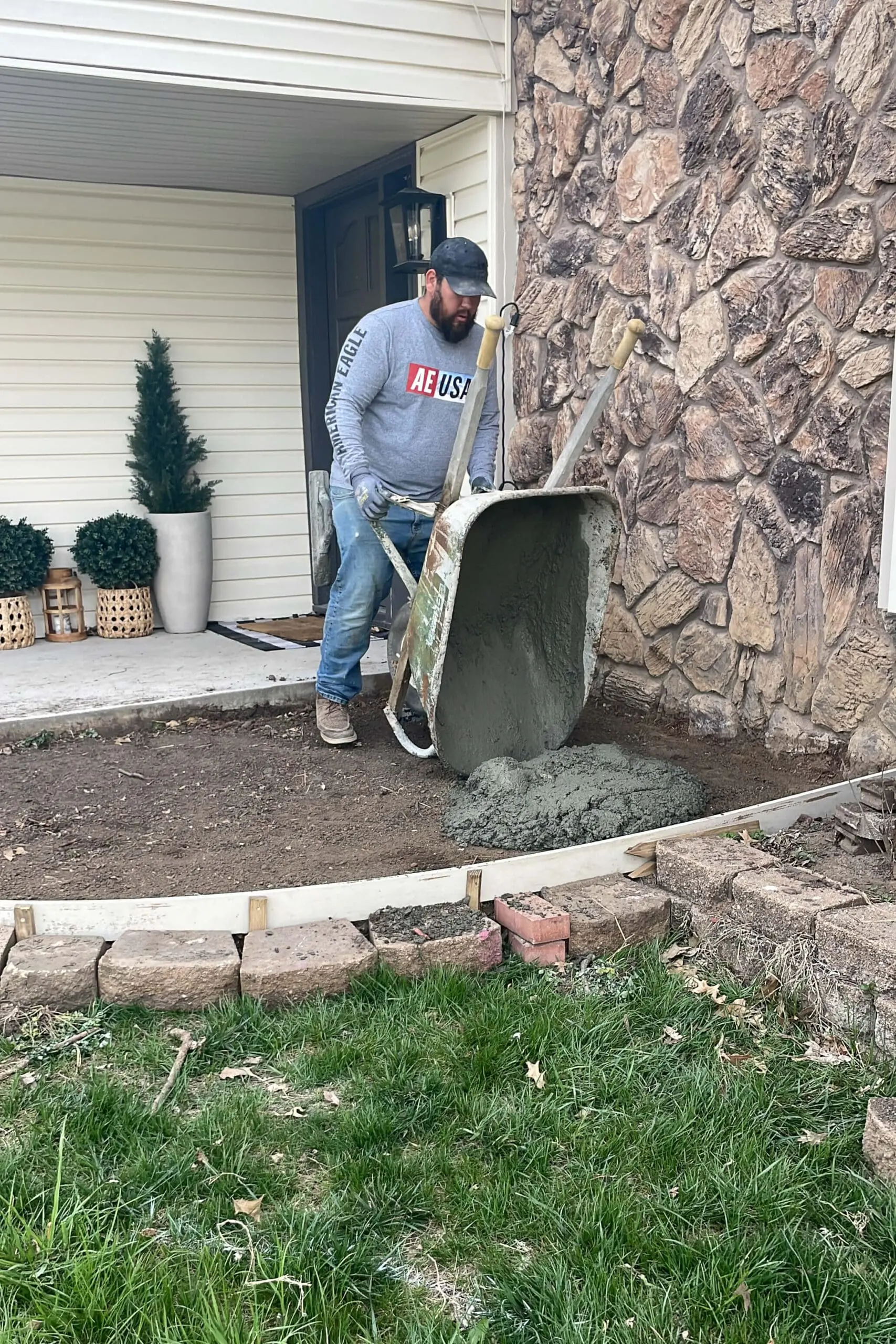 Man pouring a wheelbarrow full of wet concrete into a curved form near the front porch of a home with stone siding. The area is prepped with compacted dirt and framed with wood to shape the new sidewalk or landing.

