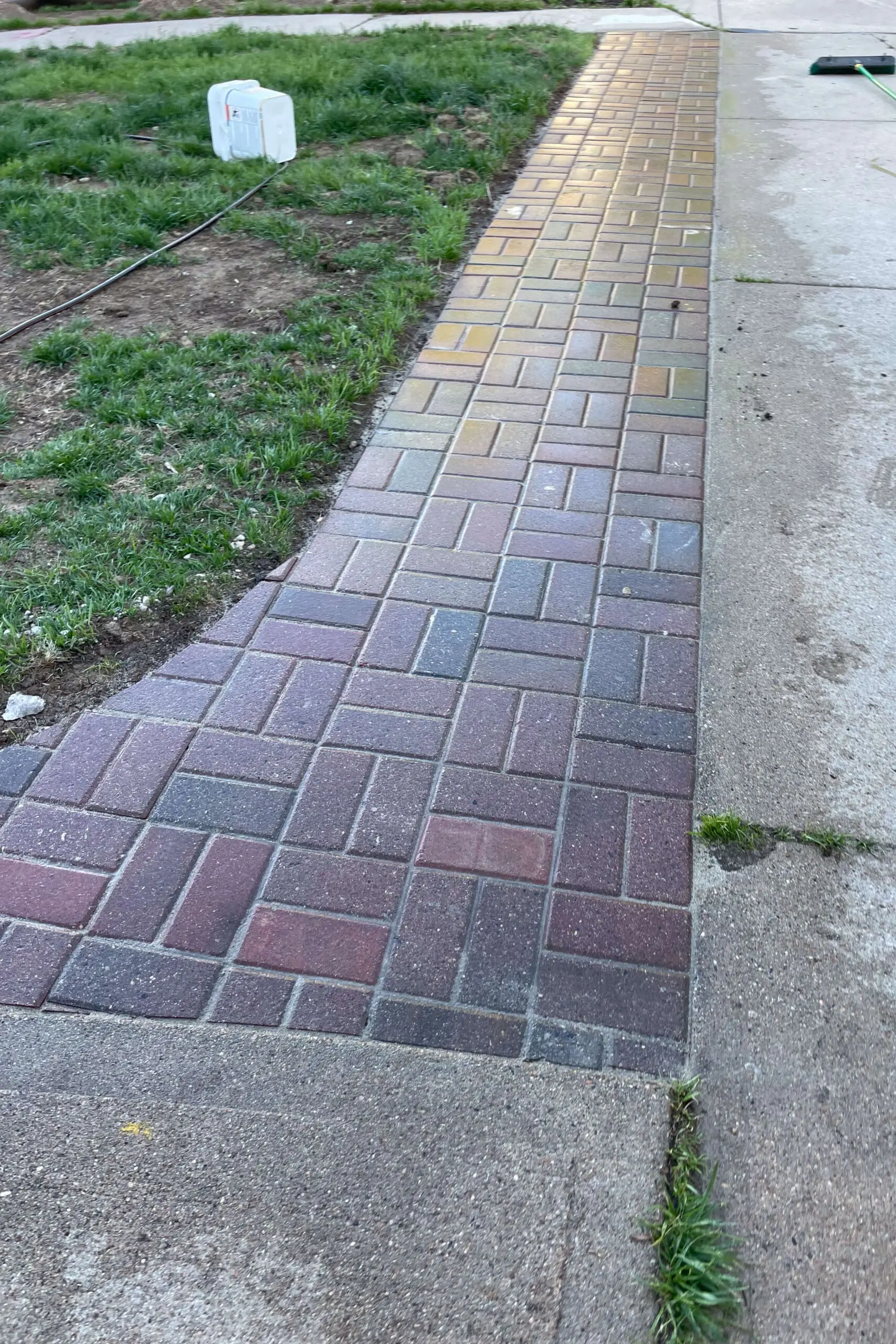 Freshly installed curved brick paver sidewalk running along the edge of a driveway as part of an exterior house remodel. The multi-colored pavers create a clean, patterned look bordered by grass and concrete, showing a major curb appeal upgrade.