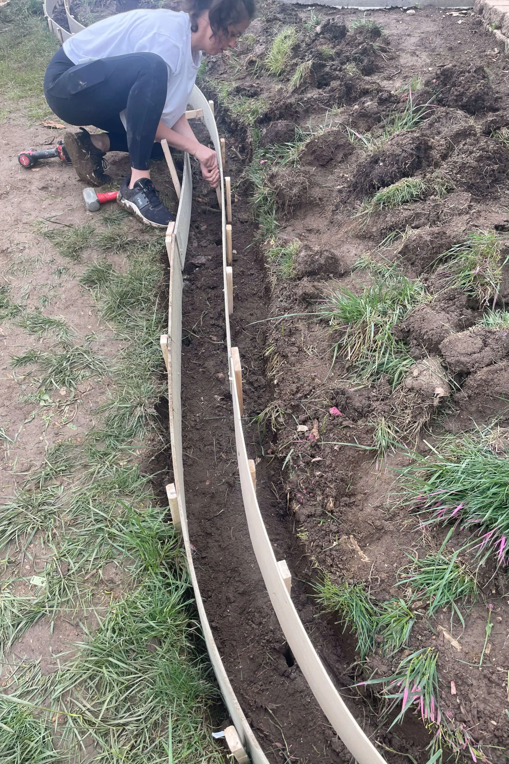 Woman kneeling in the dirt, installing flexible form boards to shape a curved concrete garden edge. Tools and disturbed grass surround the trench being prepped for pouring.