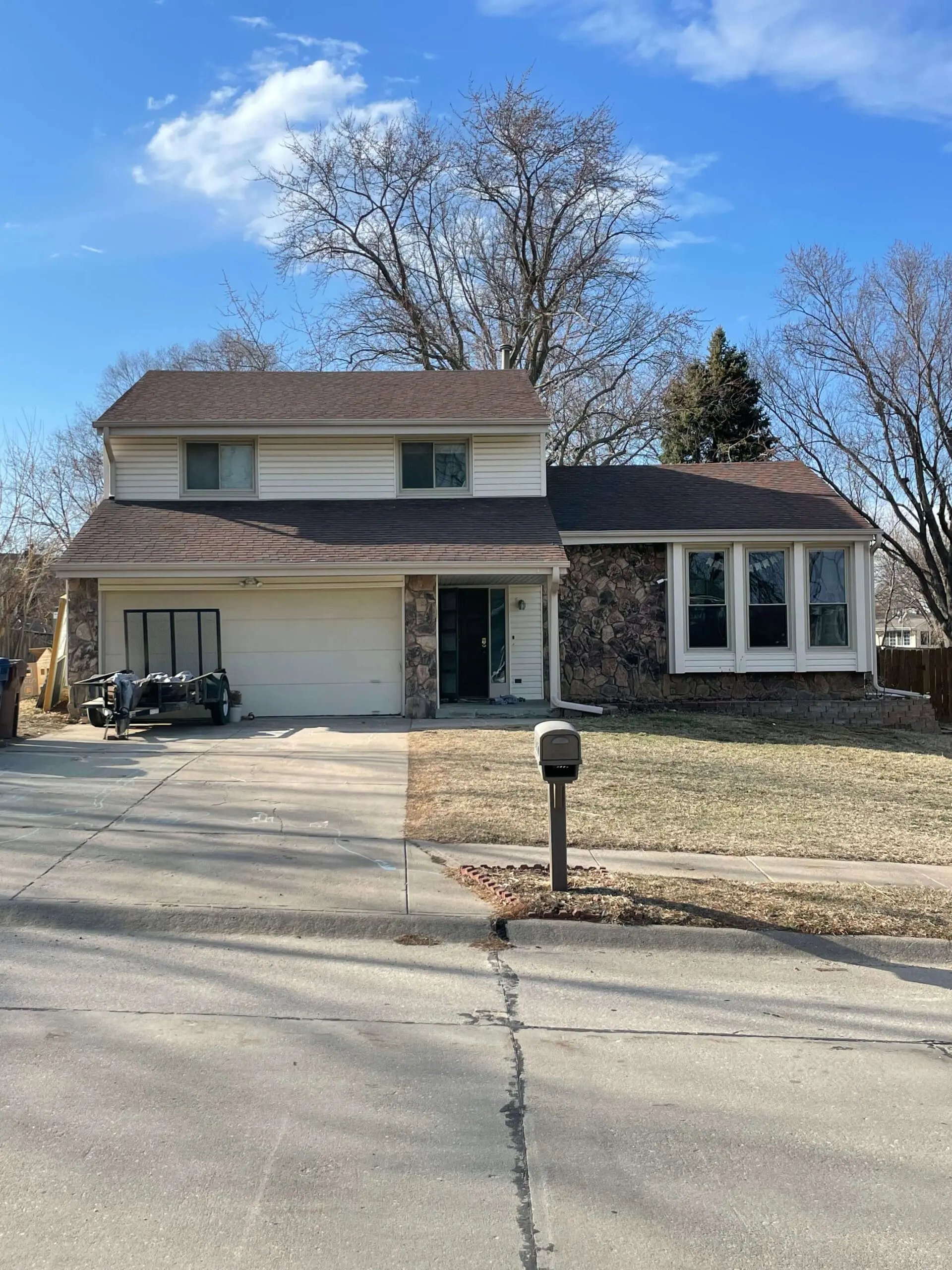 Sad and dated two-story suburban home with tan siding, stone accents, and a brown shingle roof, photographed on a clear blue-sky day before a DIY exterior house remodel. The front features a cream-colored garage door, three large front-facing windows with white trim, and a small child in a pink outfit playing near a trailer parked in the driveway. The lawn is patchy with early spring grass, and leafless trees in the background suggest late winter or early spring. 