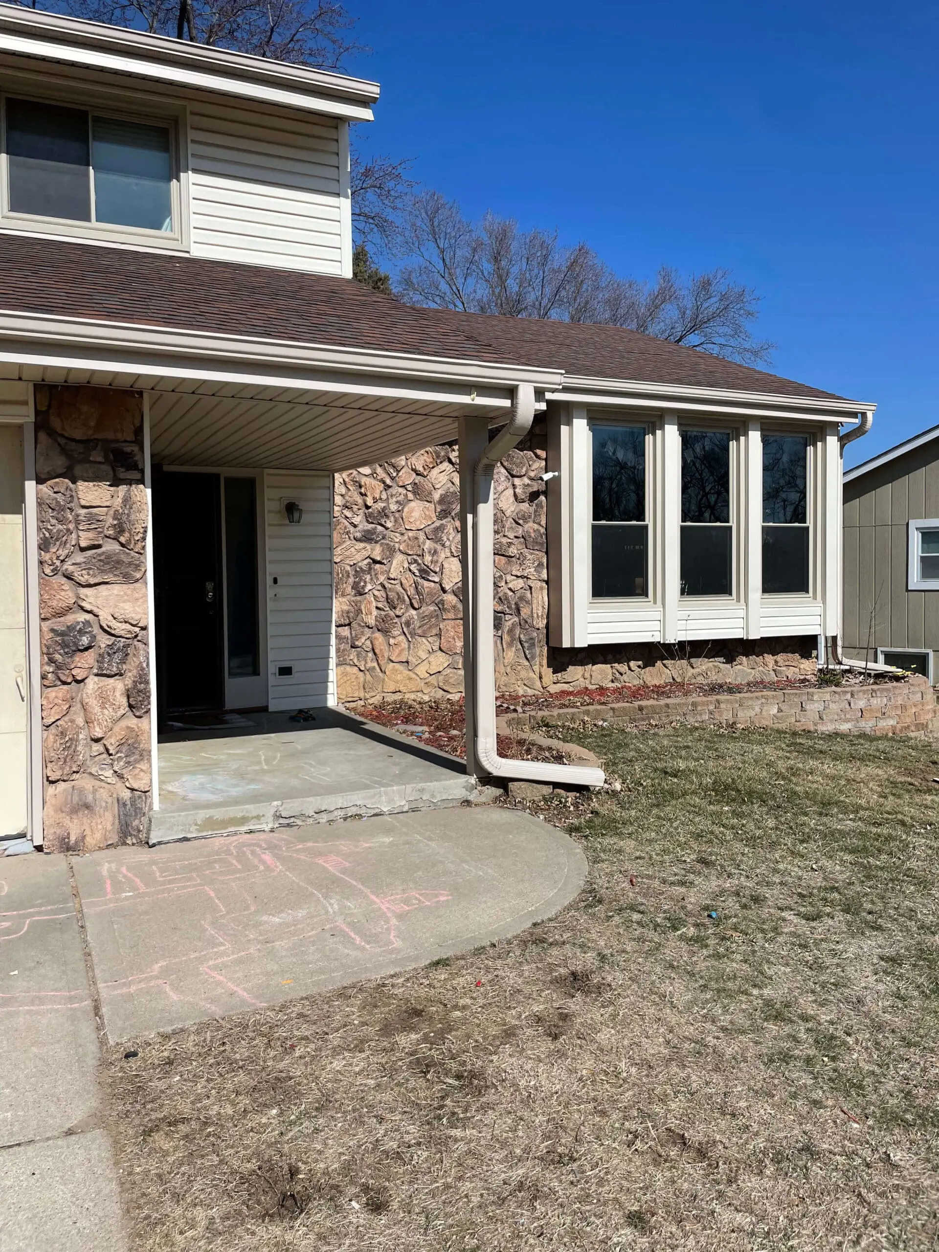 View of a home's front entry and bare yard in early spring. The stone exterior, curved concrete walkway, and sparse grass show a blank slate before landscaping or porch updates.