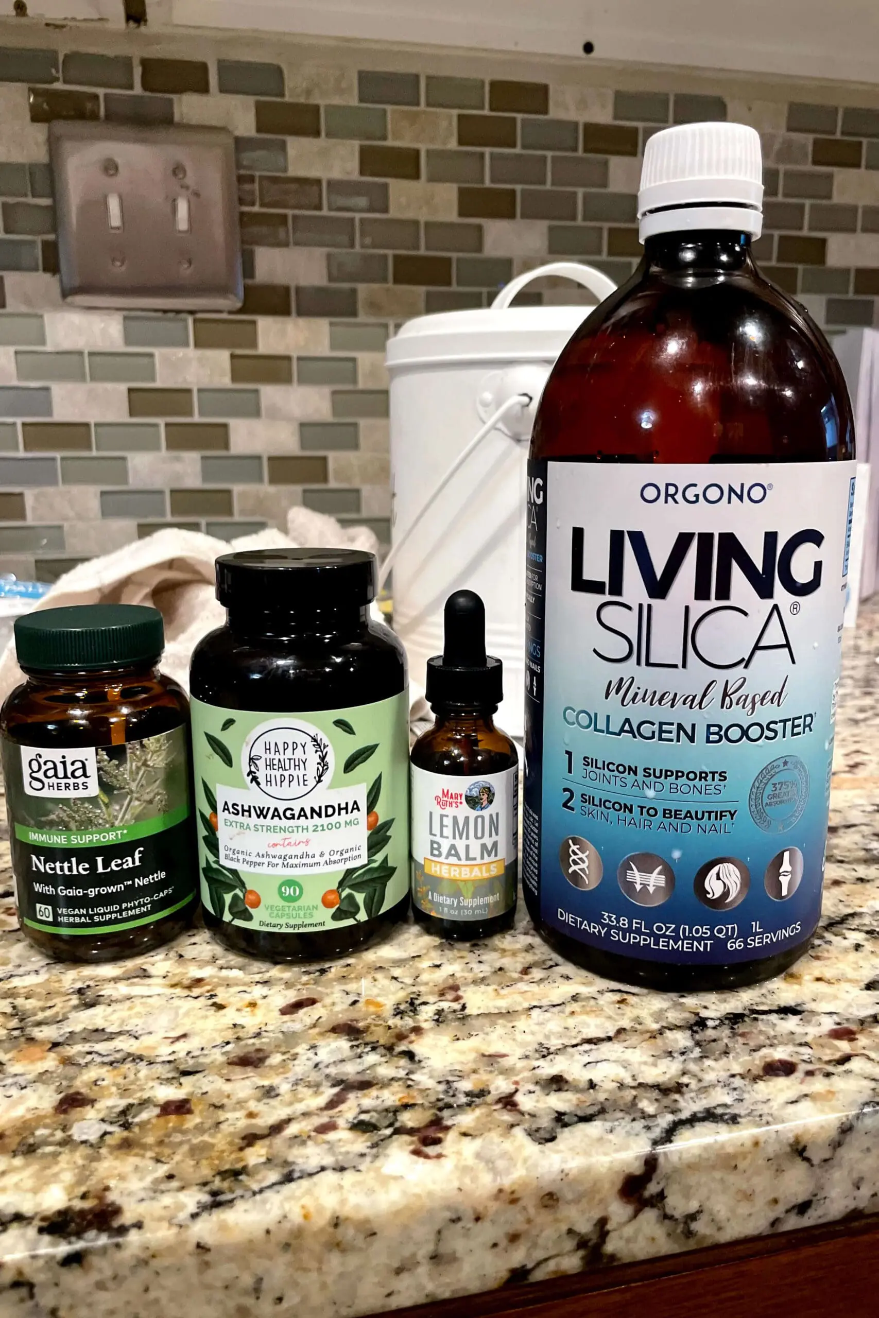 Close-up of four natural wellness products for hair health and immune support, including lemon balm, nettle leaf, ashwagandha, and silica collagen booster lined up on a kitchen counter.