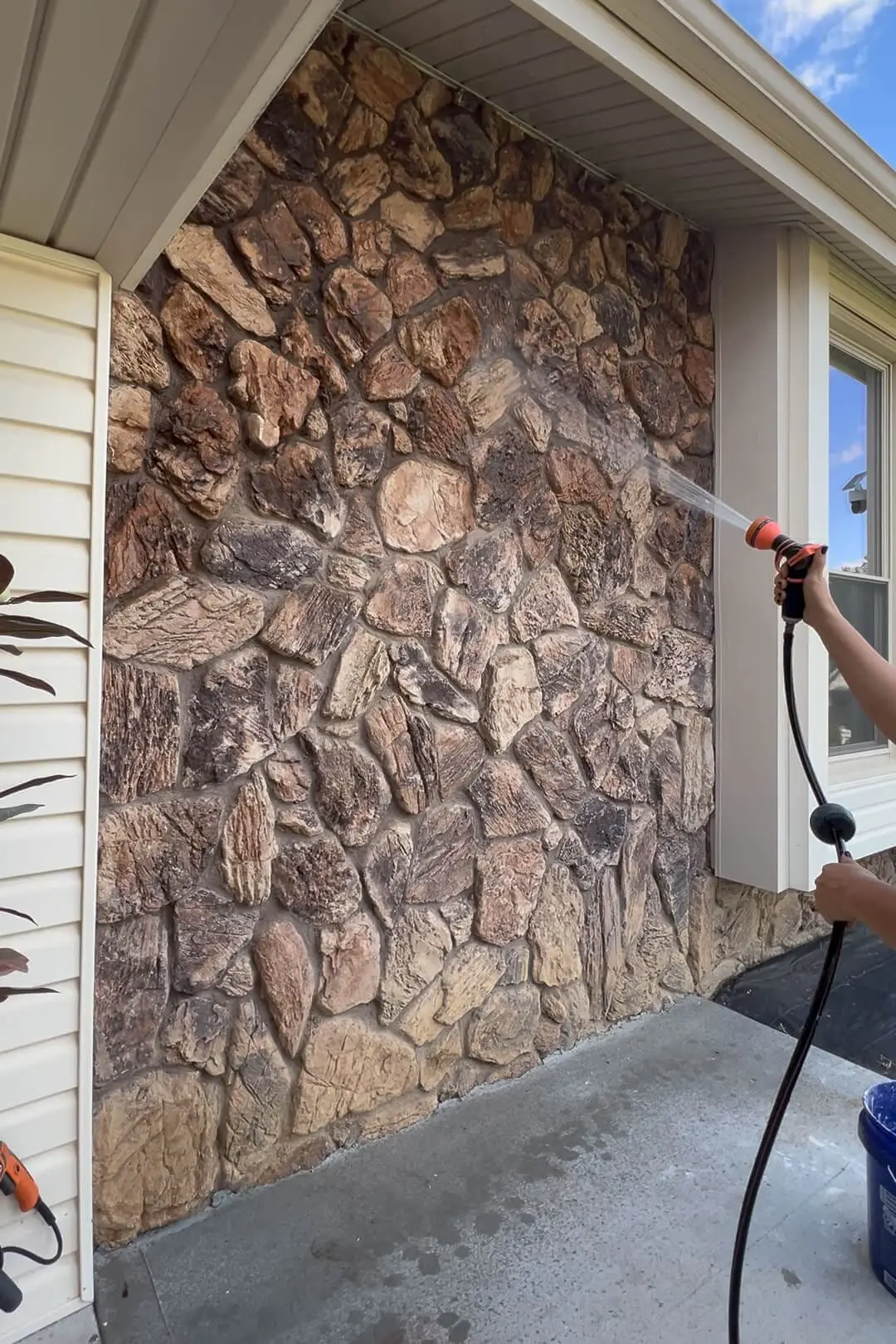Spraying down a rough brown stone wall with a garden hose before painting, part of an outdoor refresh project.