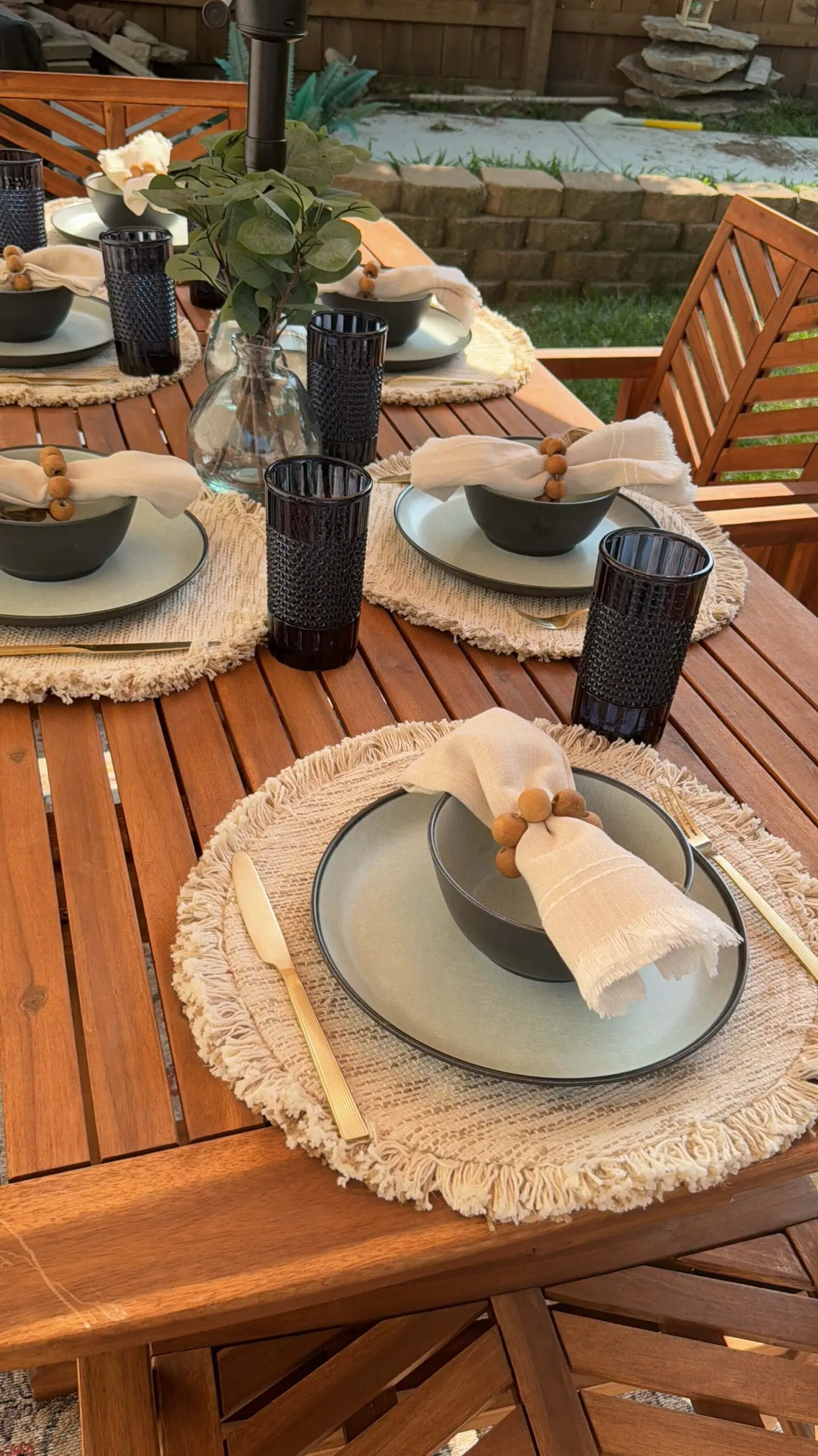 Close-up of a beautifully styled outdoor dining table with neutral place settings, gold flatware, black glasses, and fringed placemats—perfect for casual yet elegant outdoor entertaining.
