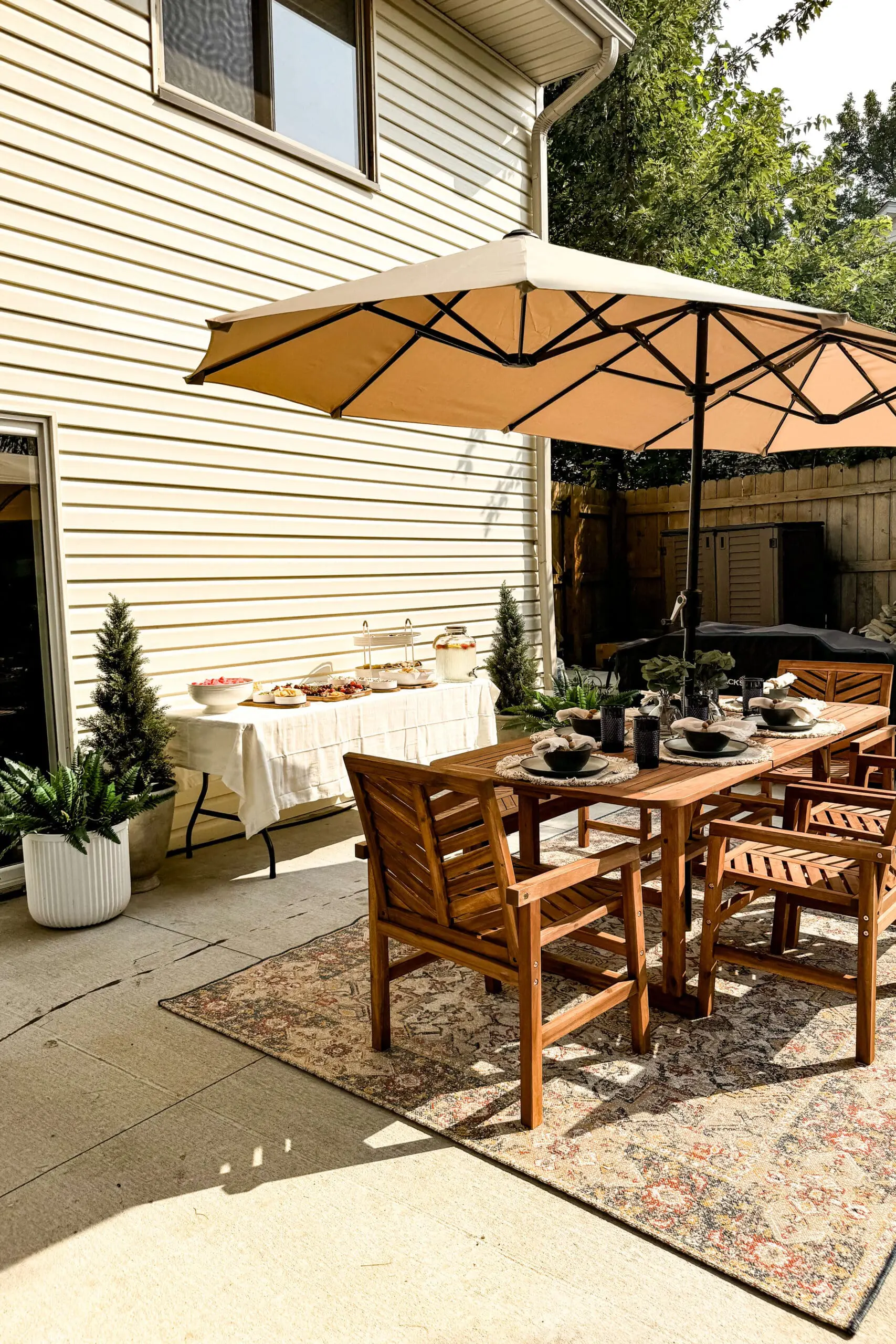 Outdoor wooden dining table setup under a large patio umbrella, styled for summer backyard entertaining with a nearby buffet table.