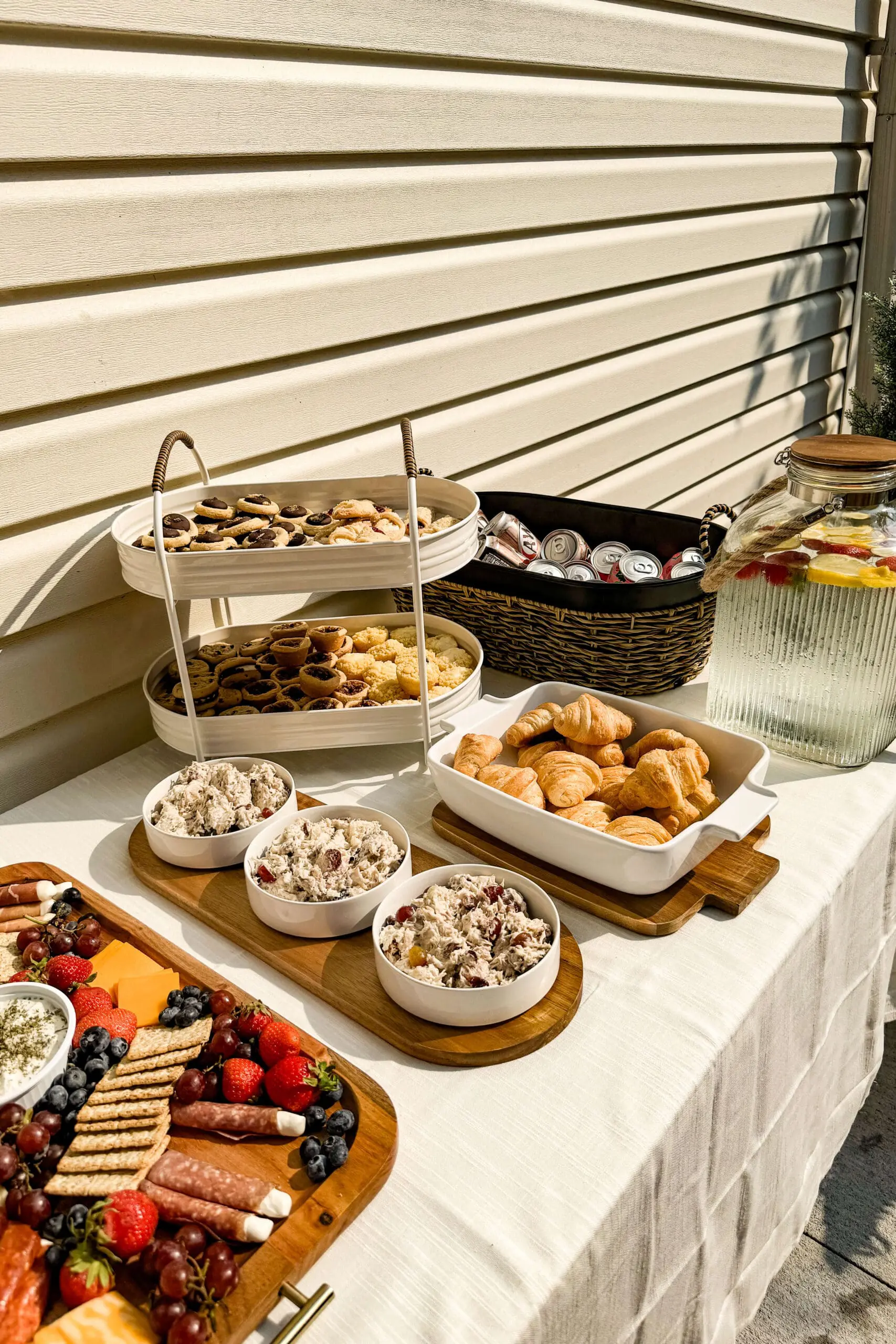 Assorted mini pastries, chicken salad, cookies, and fruit water on a backyard brunch table—easy hosting ideas for outdoor gatherings.

