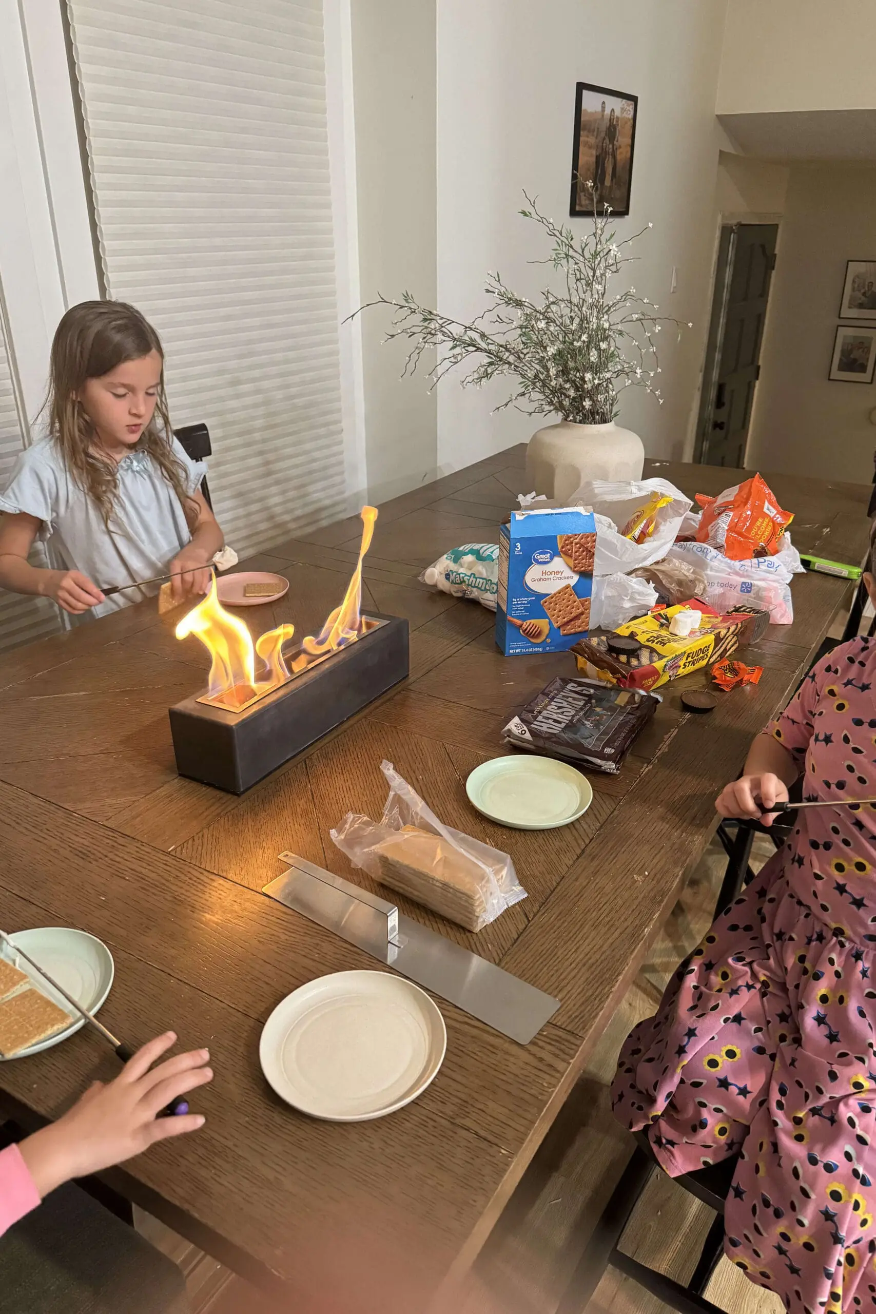 Children gathered around a tabletop fire pit indoors, roasting marshmallows and prepping s’mores—perfect for year-round hosting ideas.