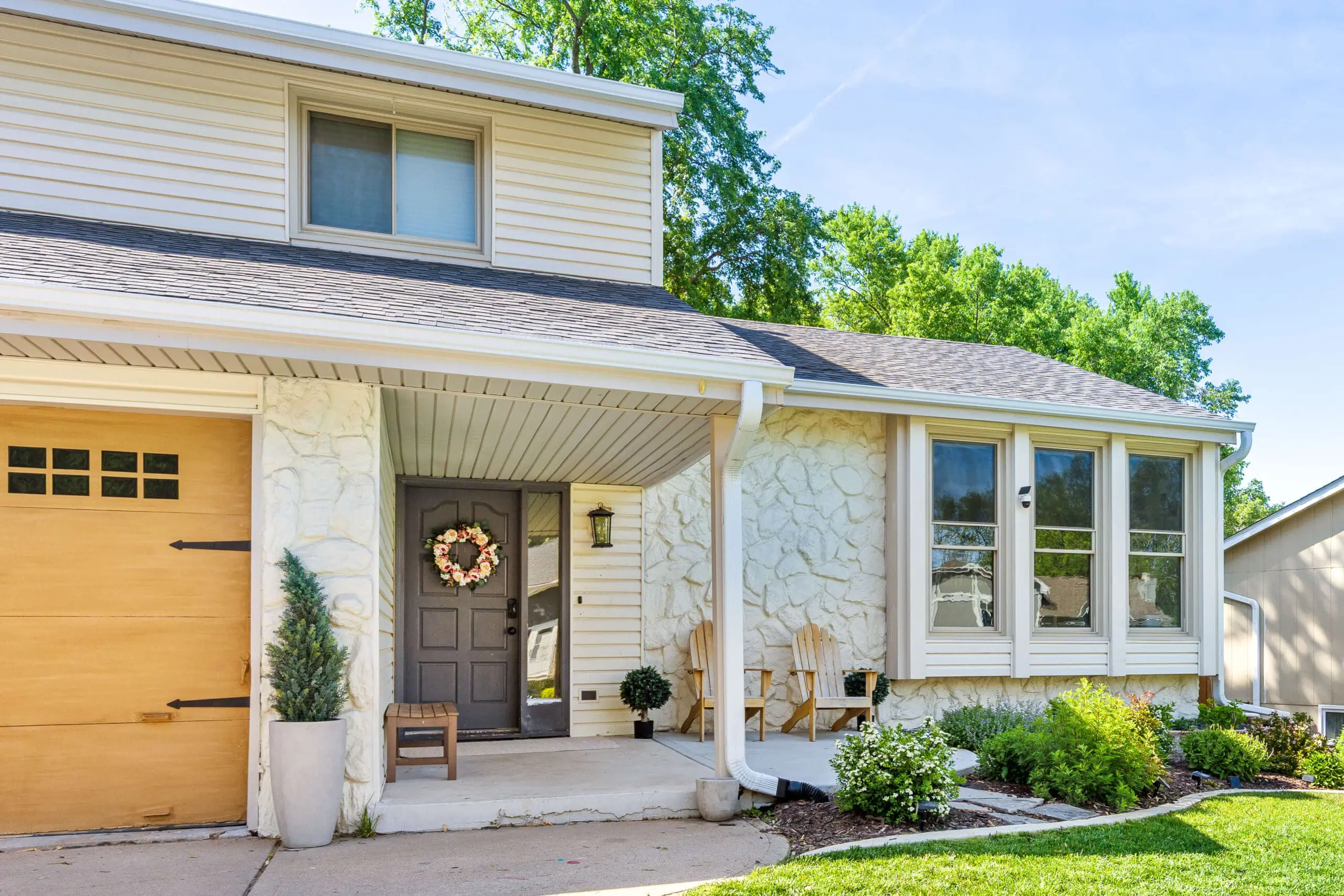 Close-up of a cozy front porch with a dark painted door, floral wreath, textured stone siding, and natural wood Adirondack chairs surrounded by blooming landscaping.