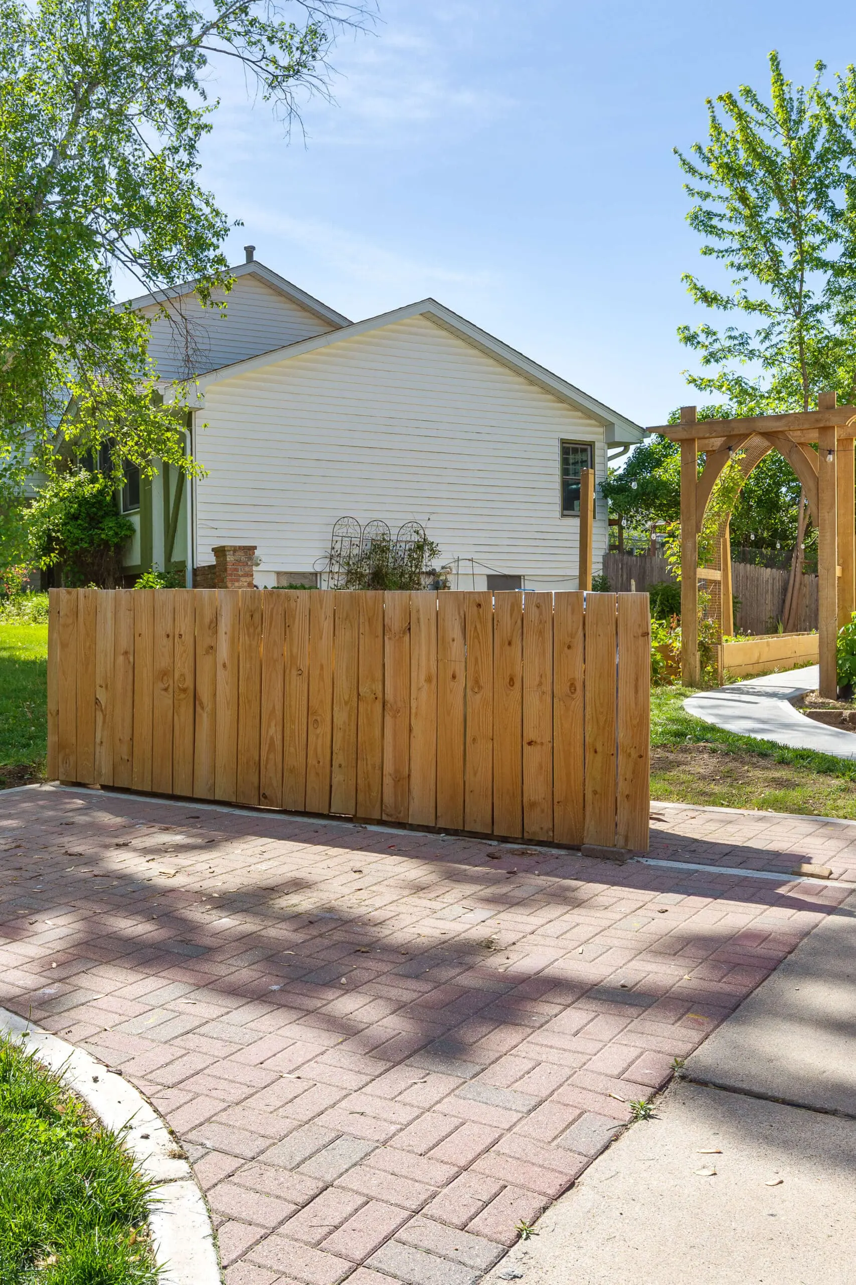 DIY wooden fence enclosure built to hide trash cans, set on a red paver pad with a curved sidewalk and a wooden garden pergola nearby.