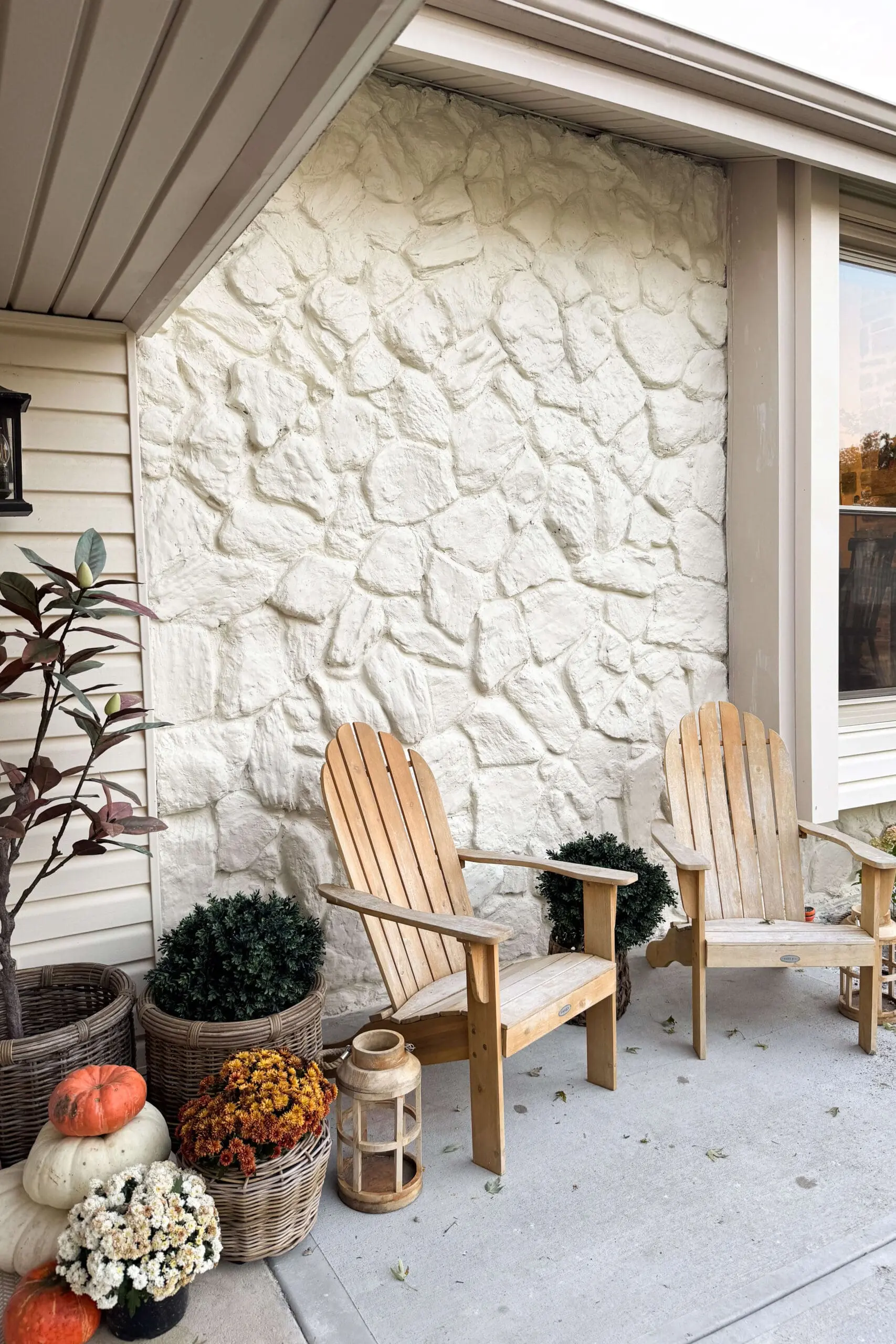 Painted white stone wall with a cozy porch setup featuring wooden Adirondack chairs, mums, pumpkins, and wicker baskets.