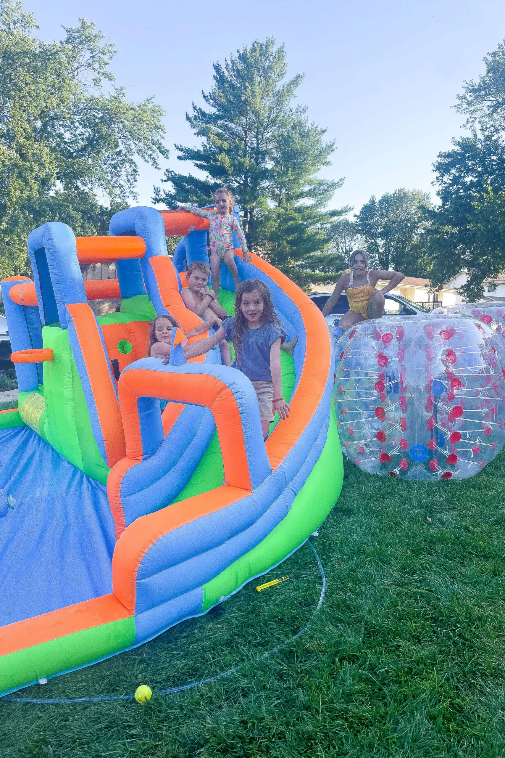 Group of kids smiling and playing on a colorful inflatable water slide and bounce structure, with a bumper ball nearby—backyard party fun and outdoor entertainment for families.