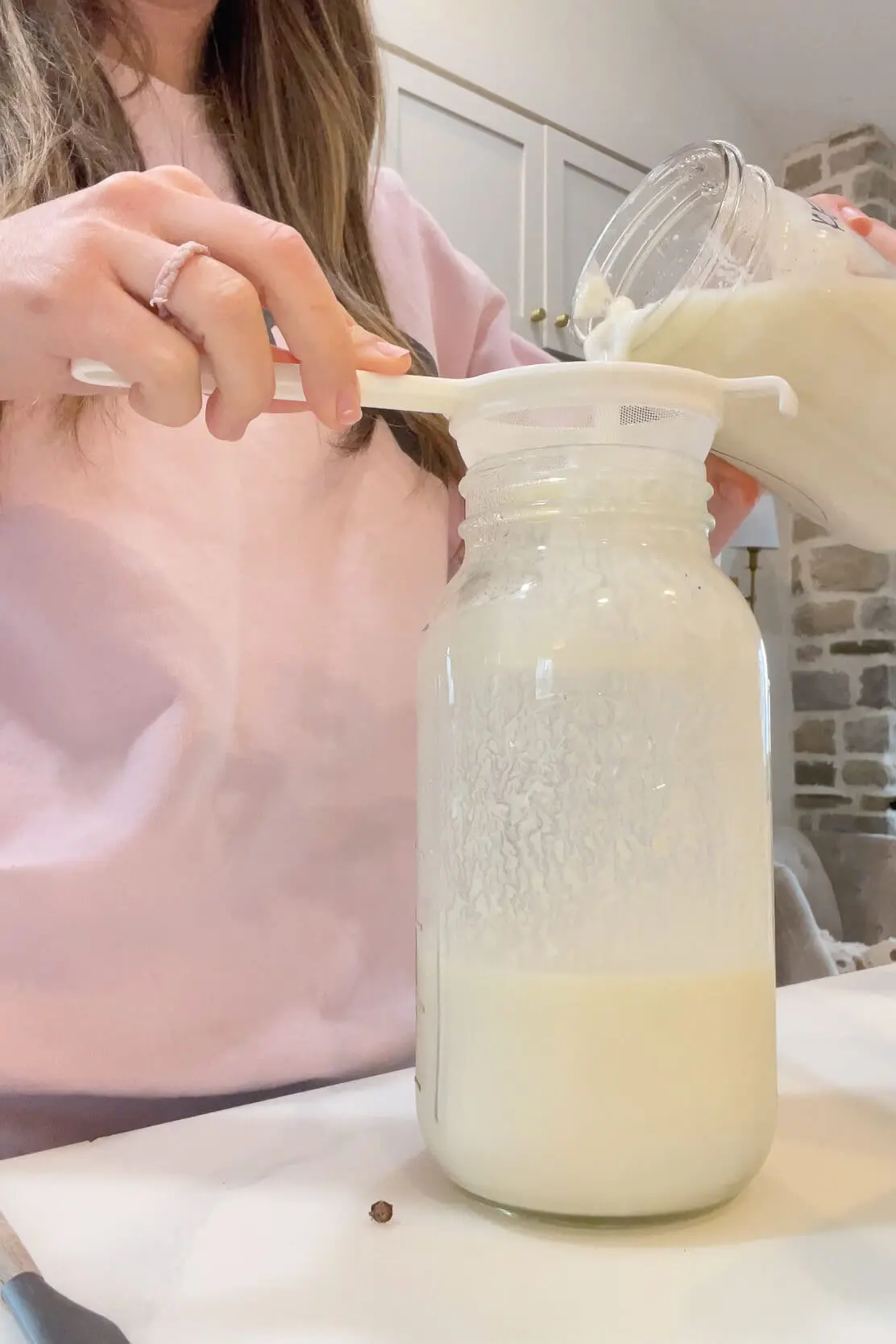 Pouring freshly fermented milk kefir through a plastic mesh strainer into a clean jar while showing how to make milk kefir at home.