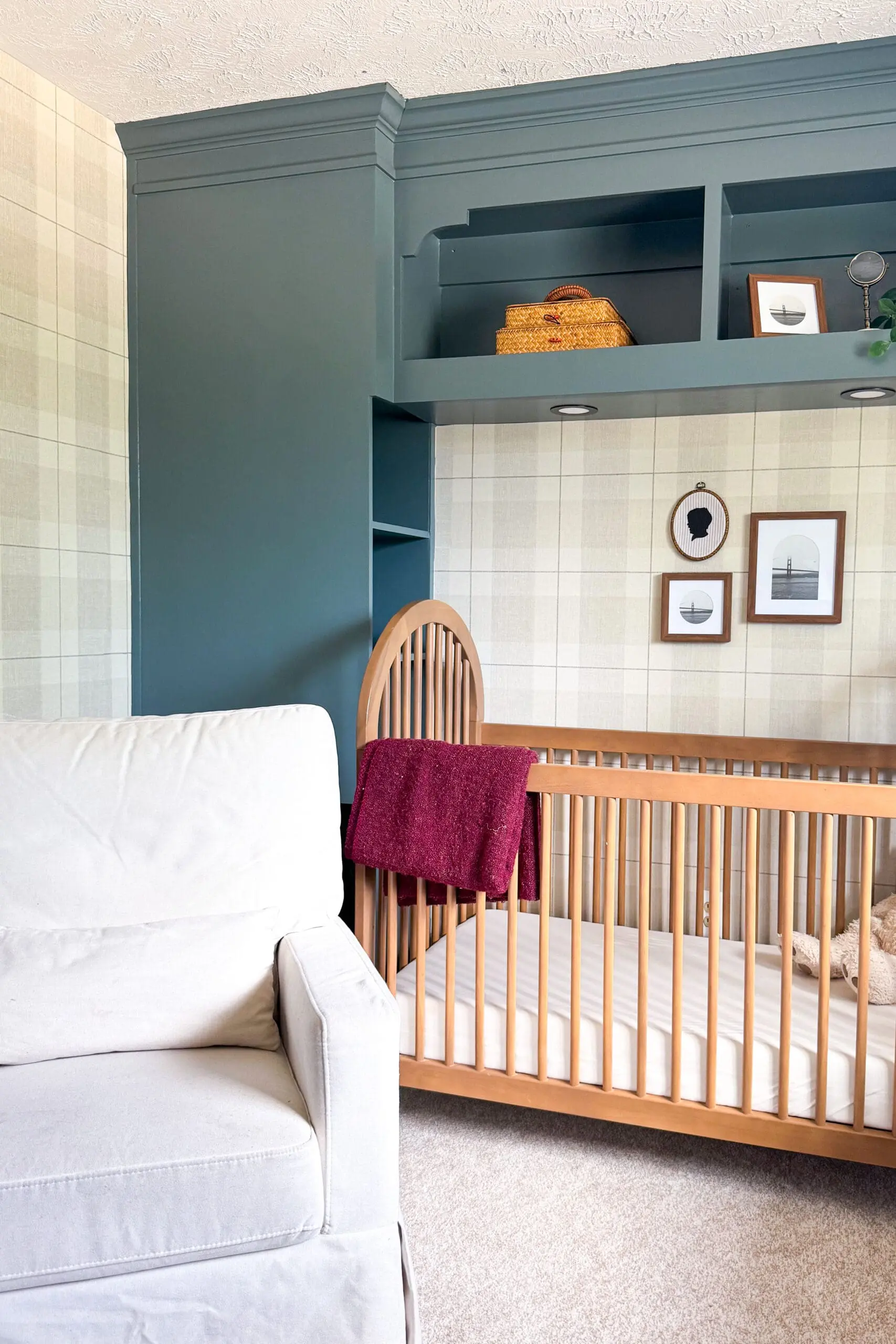 Side angle of crib in a nursery with plaid wallpaper and deep green built-in shelves holding decorative accents and a woven basket.