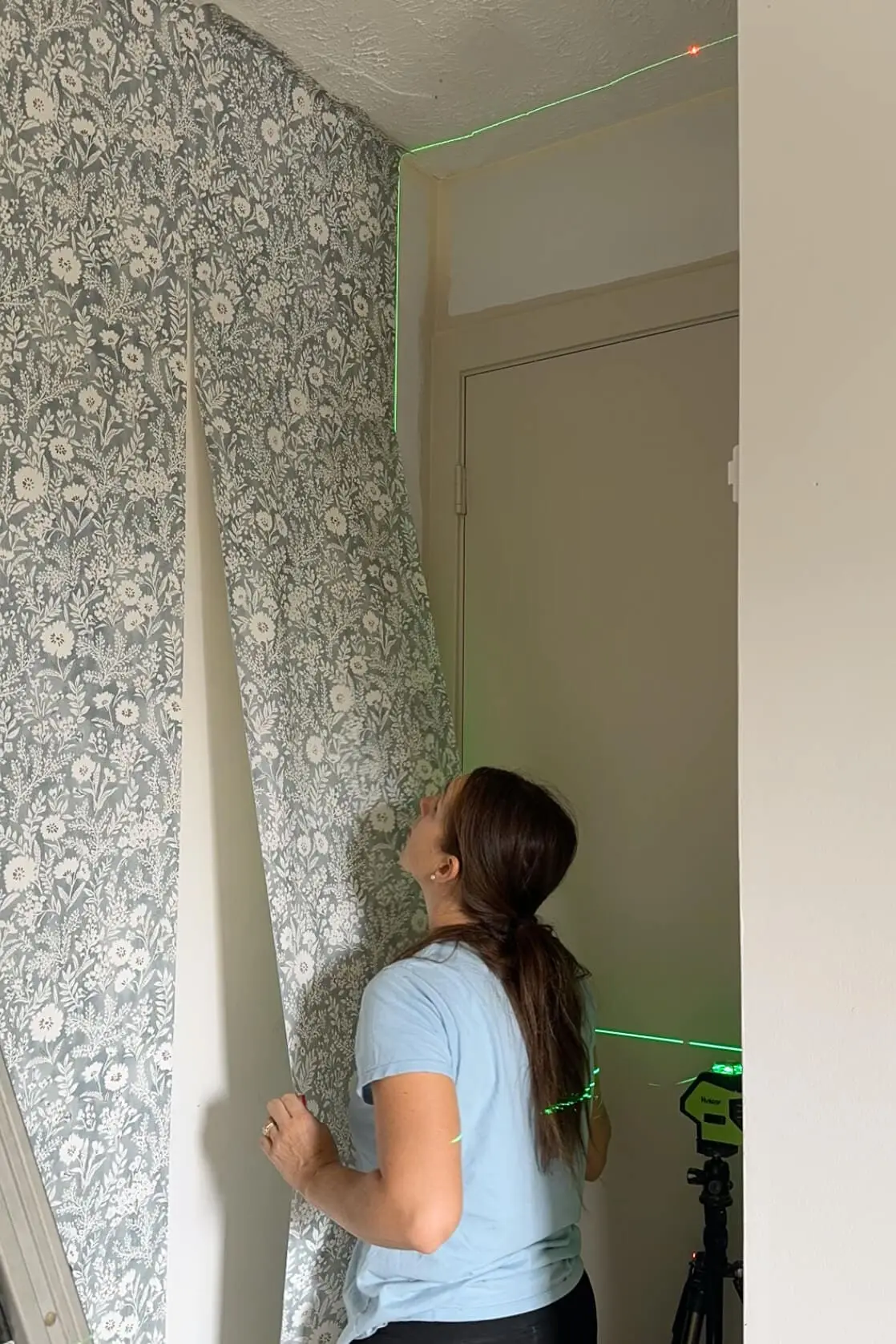 Woman aligning the next panel of floral wallpaper along the ceiling edge using a green laser level for precision.
