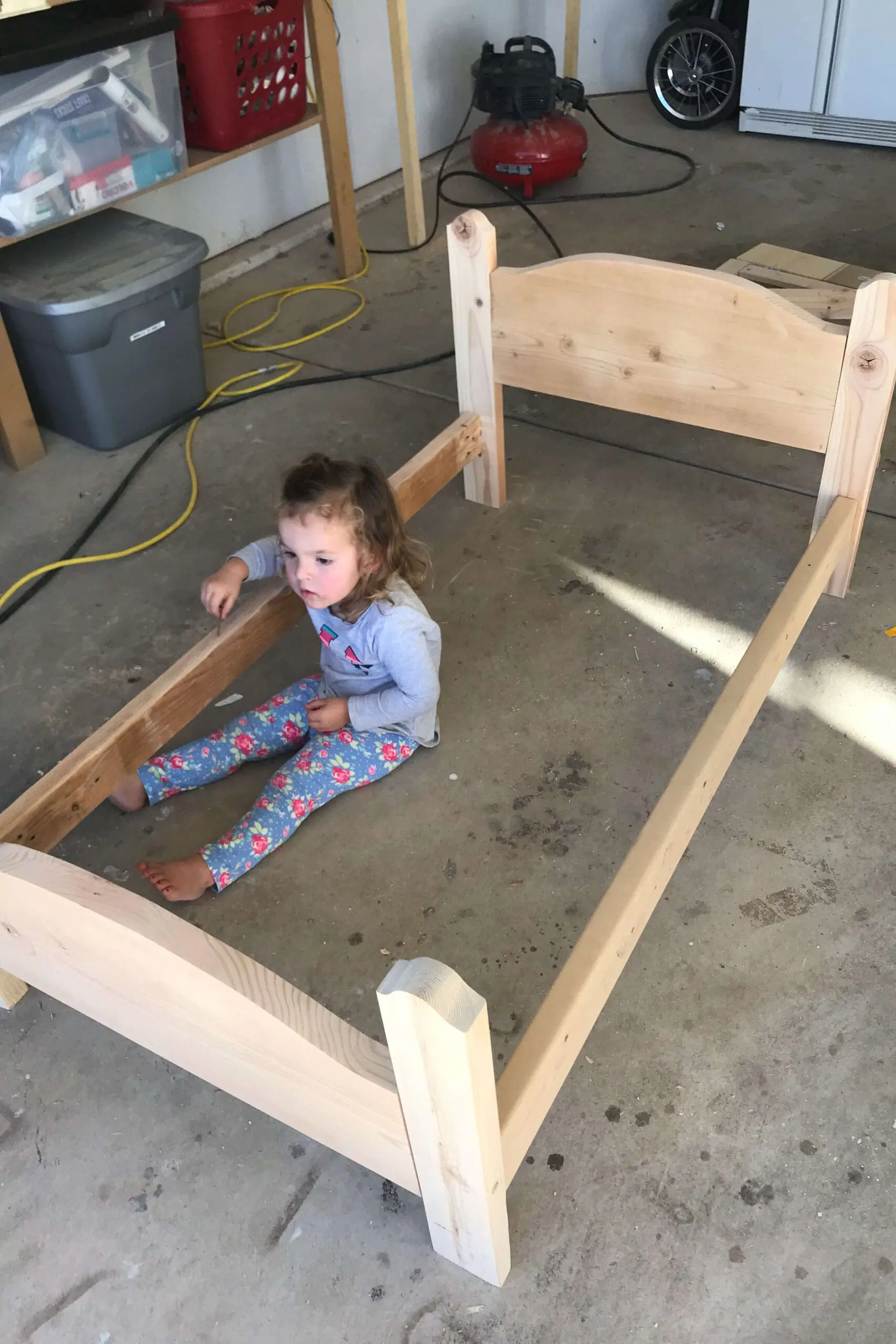 Partially assembled wooden bed frame in a garage with a young child sitting inside, helping during the DIY build process.