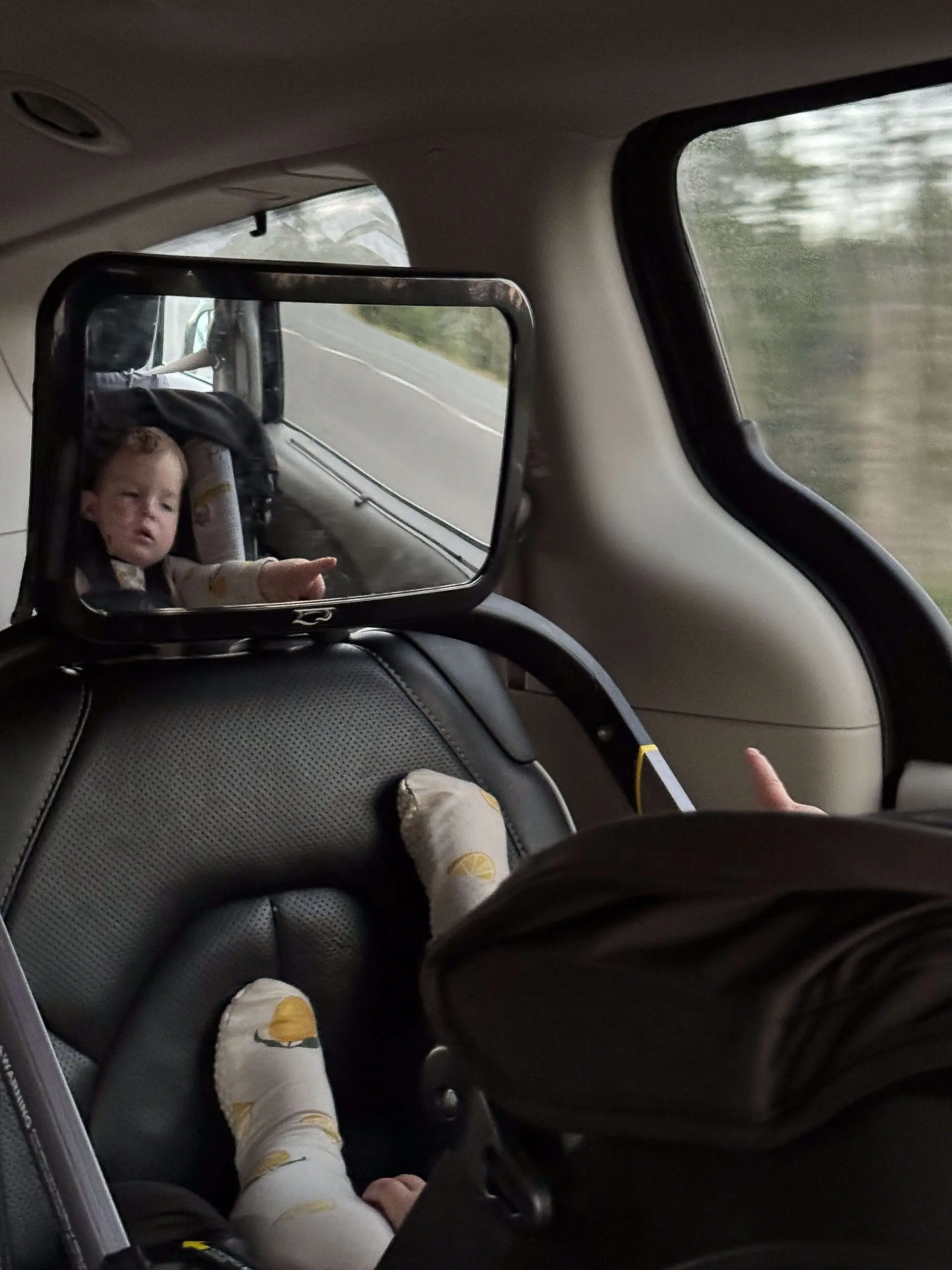 Rear-facing baby seen through a backseat mirror during a car ride, with little feet propped up and a peaceful road trip backdrop outside the window.