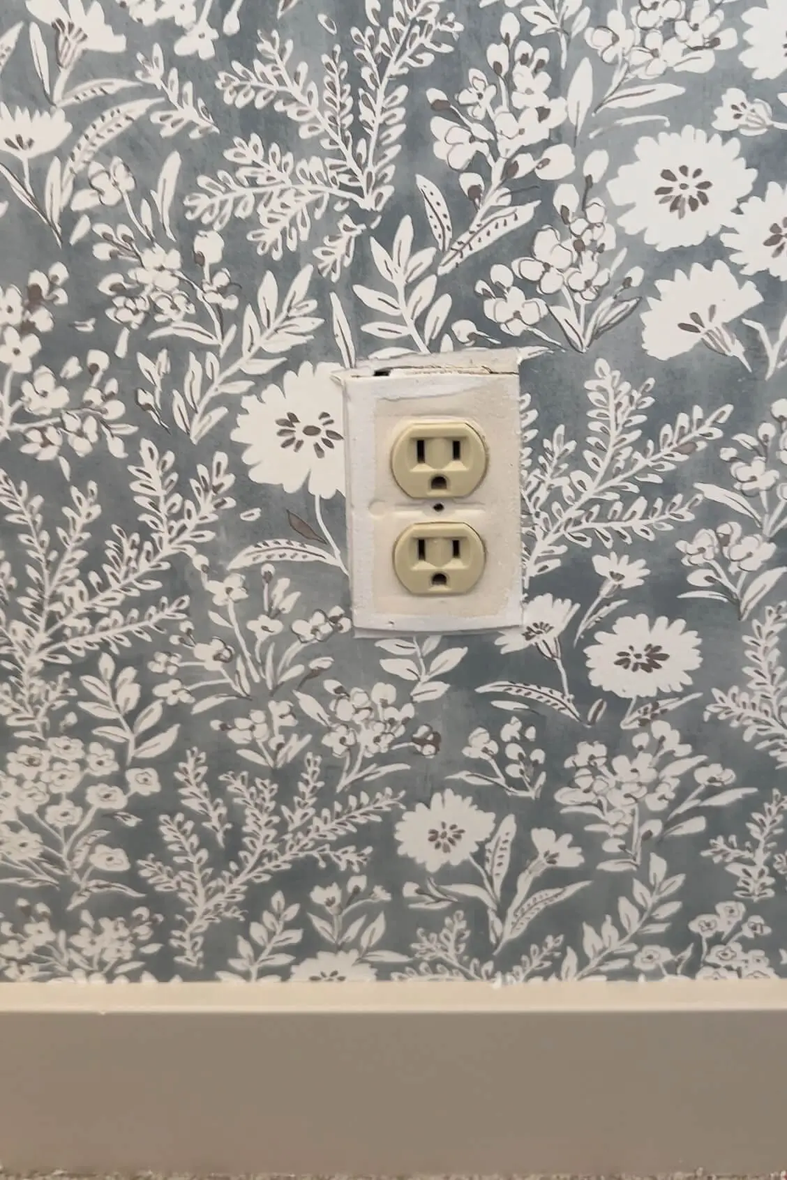 Standard white outlet on a blue floral wallpapered wall, showing the unfinished look before adding a camouflaged cover.