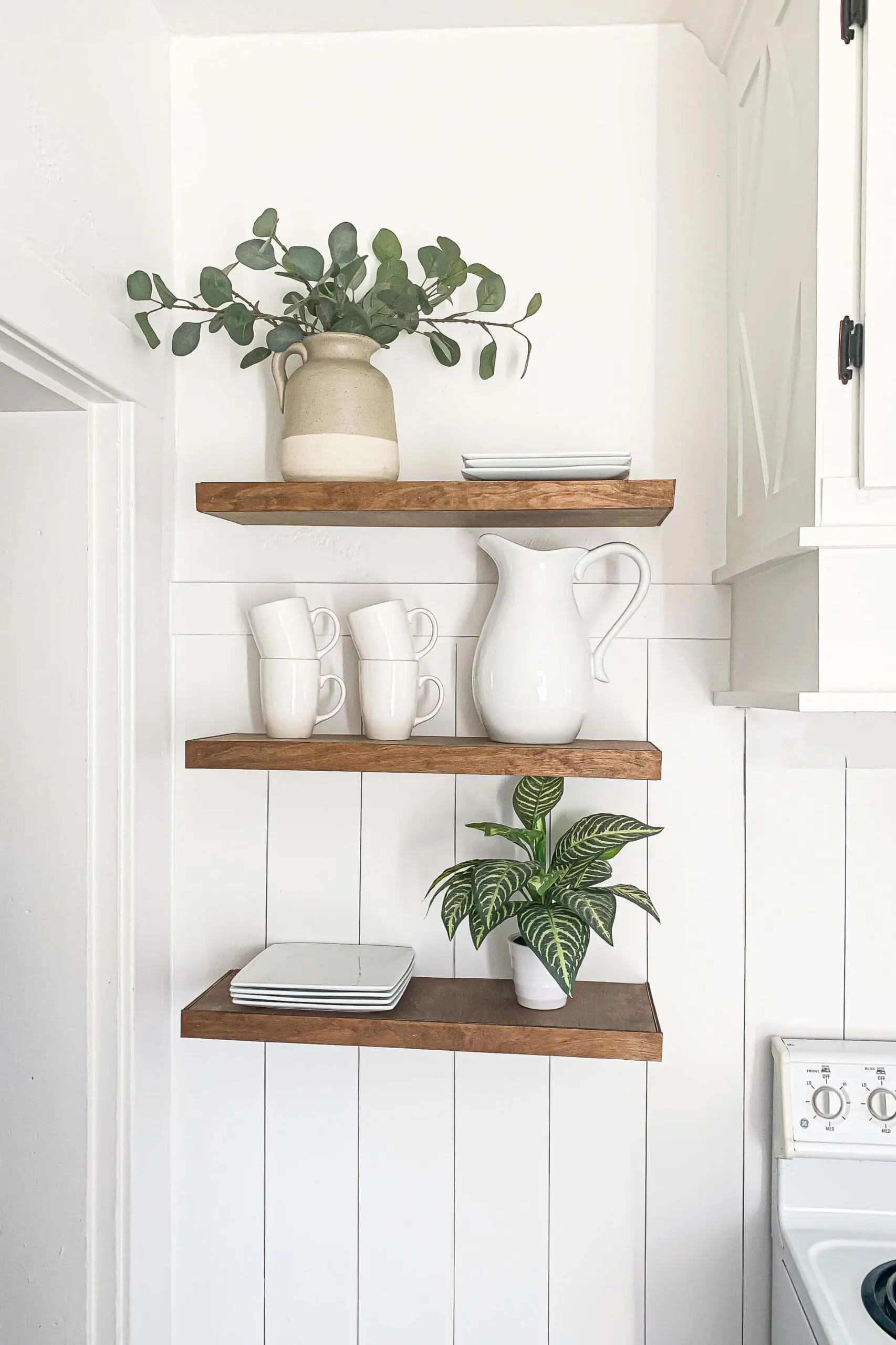 Straight-on view of handmade DIY floating shelves installed without brackets, featuring wood tones against a white kitchen wall, styled with dishes and a ceramic jug for a clean, modern farmhouse look.