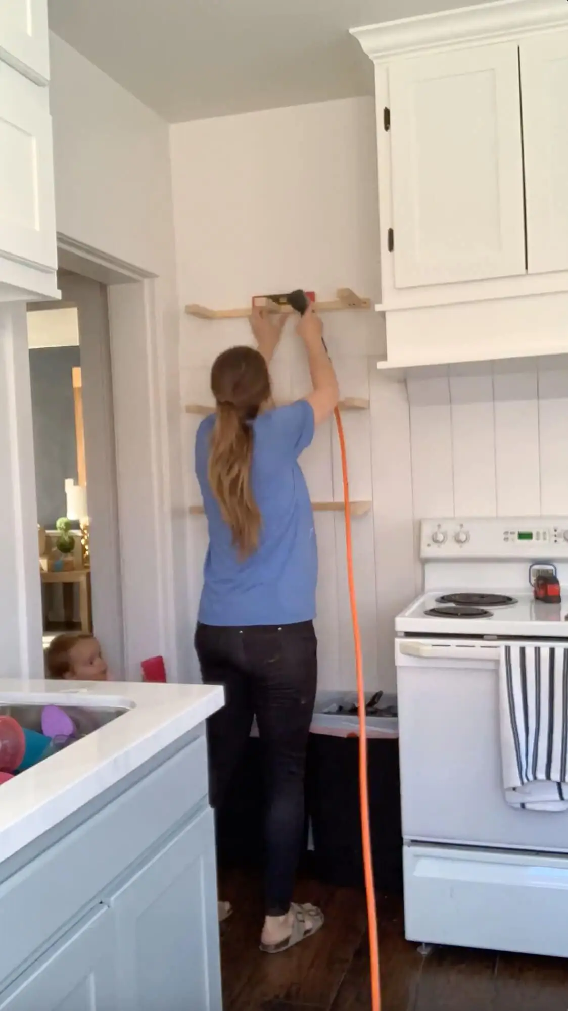 Using a brad nailer to secure the top cleat for a floating shelf, with all three shelf supports aligned on a white shiplap kitchen wall during a home DIY build.