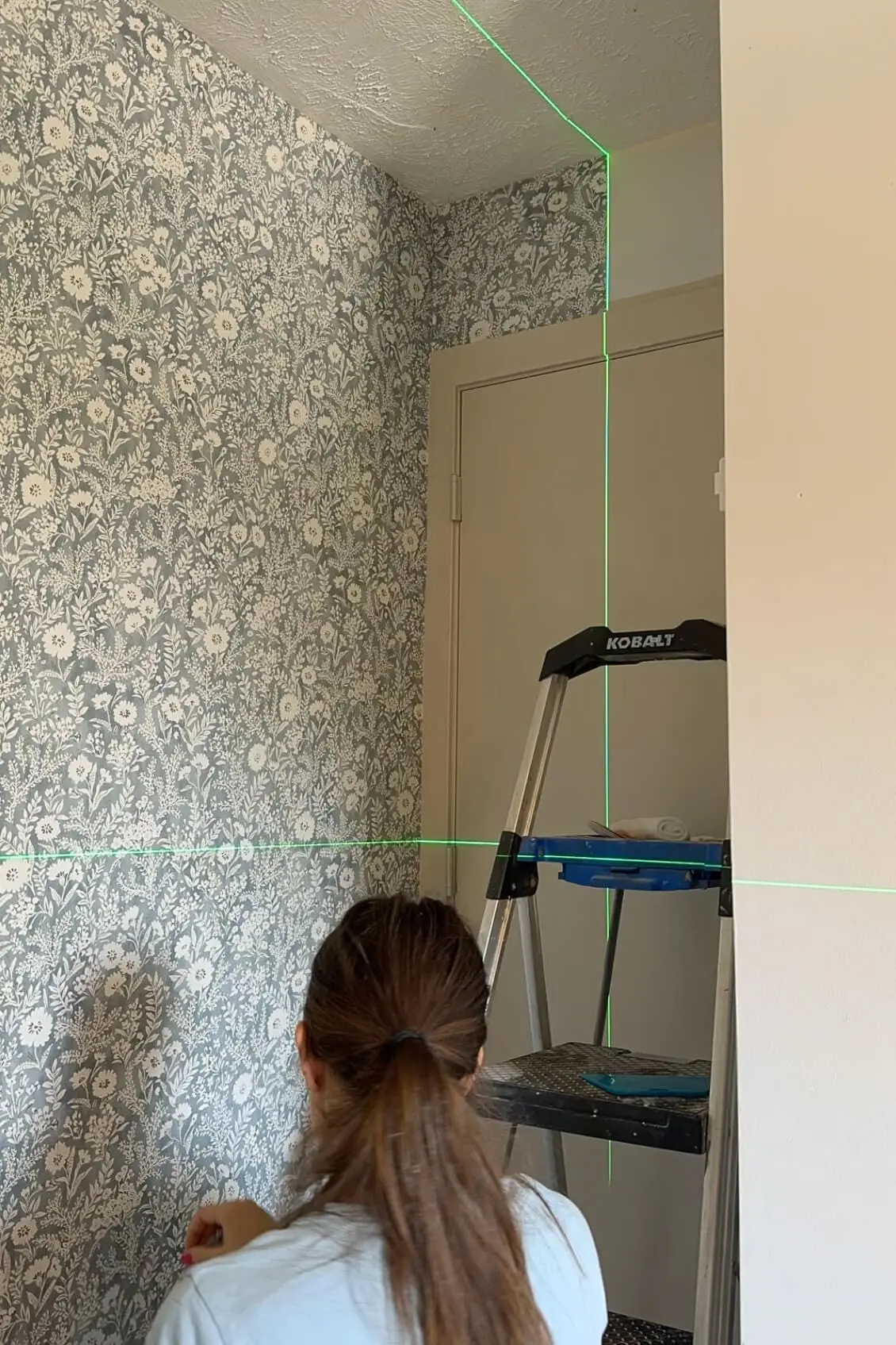 Woman reviewing wallpaper alignment with a laser level, standing in front of a nearly completed floral wallpapered wall.