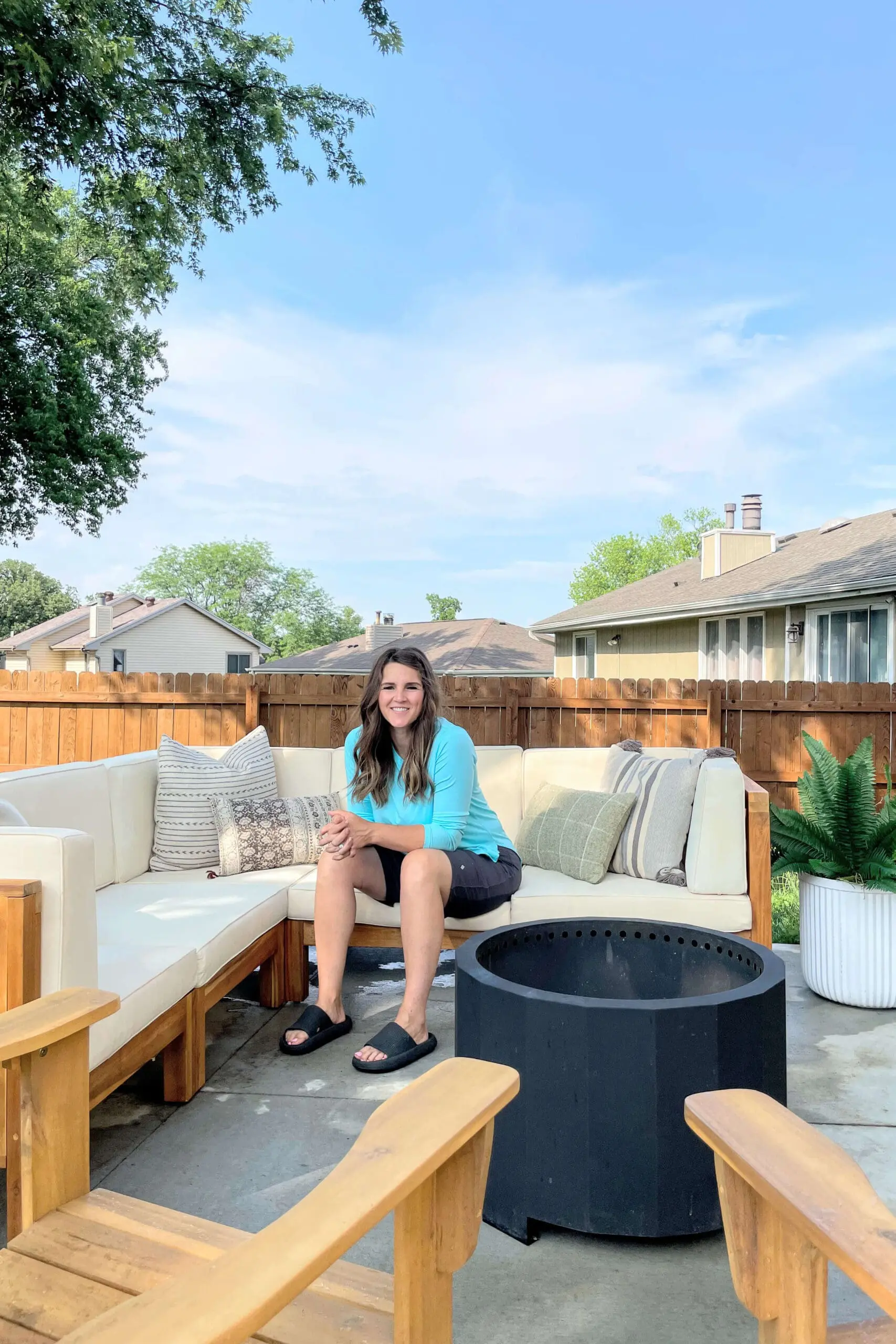 Woman sitting on a clean concrete patio with a wood and cream outdoor sectional, black fire pit, and decorative pillows