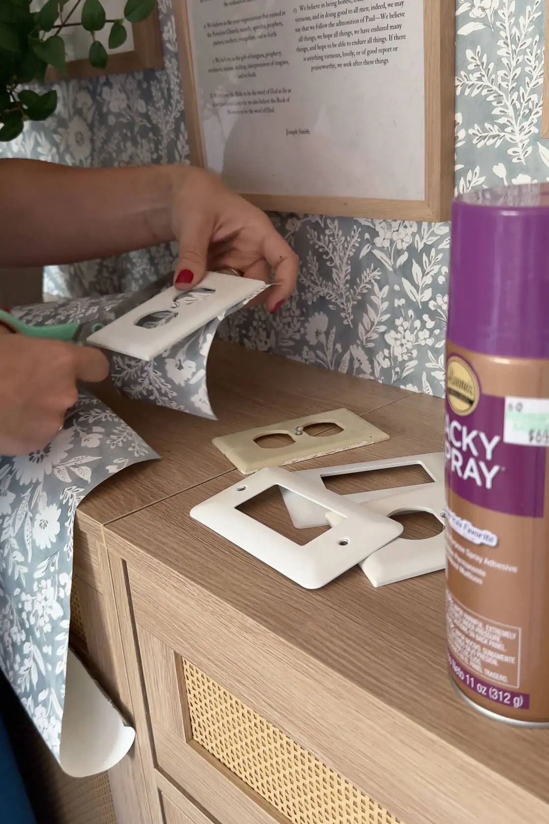 Hands cutting floral wallpaper to size around a white wall plate, mid-DIY project.