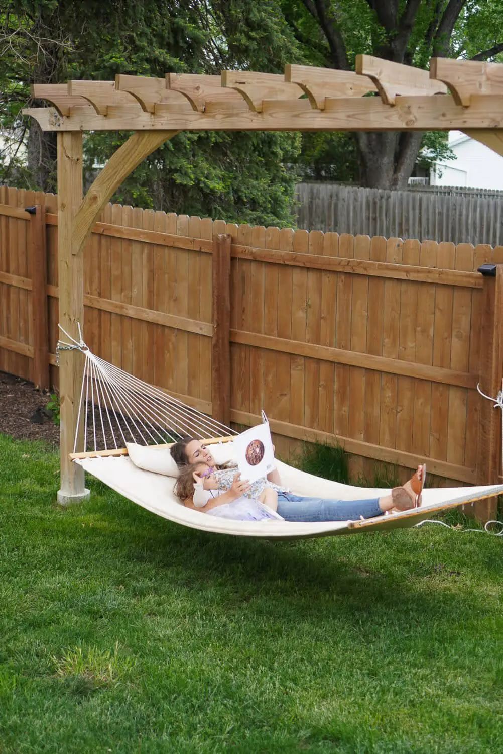 Mom and daughter relaxing together in a hammock under a wooden pergola, reading a picture book in a sunny backyard.